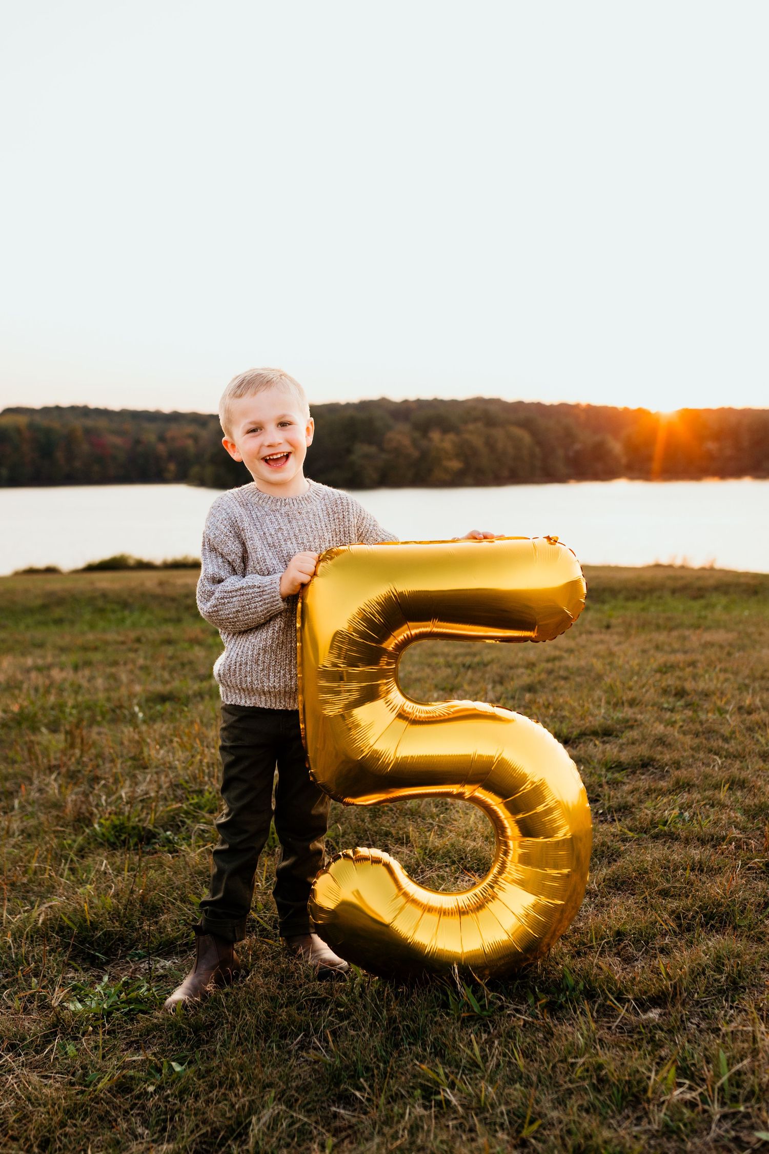 Happy boy holding a large gold number 5 balloon at sunset by a lake.