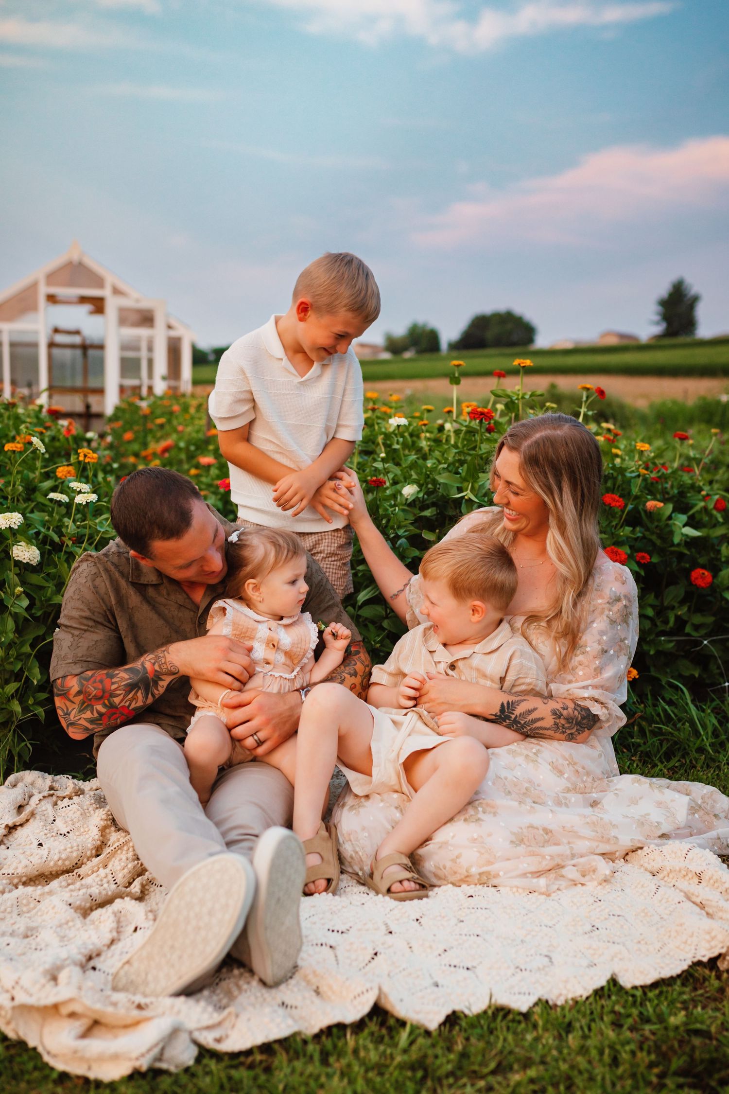 Family of five sitting on a blanket in a flower garden at sunset, with two toddlers and an older boy sharing a joyful moment.