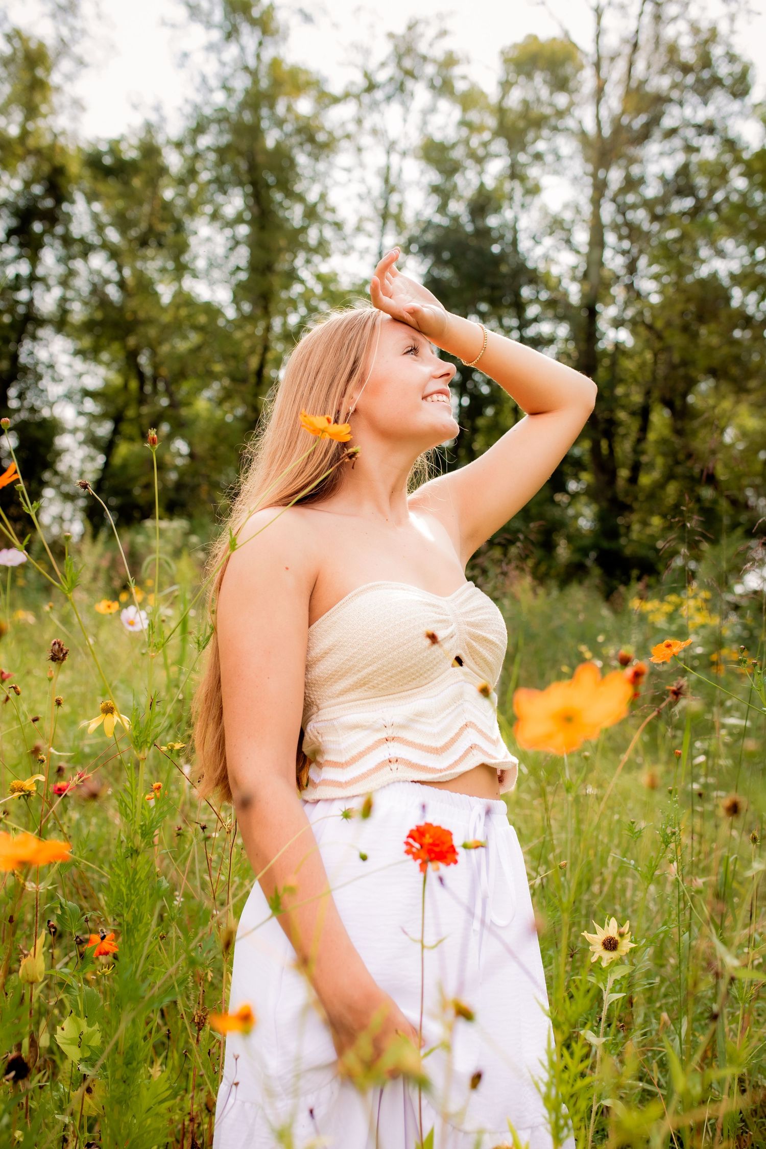 Blonde teen girl in cream crop top and white skirt poses among colorful wildflowers in sunny field.