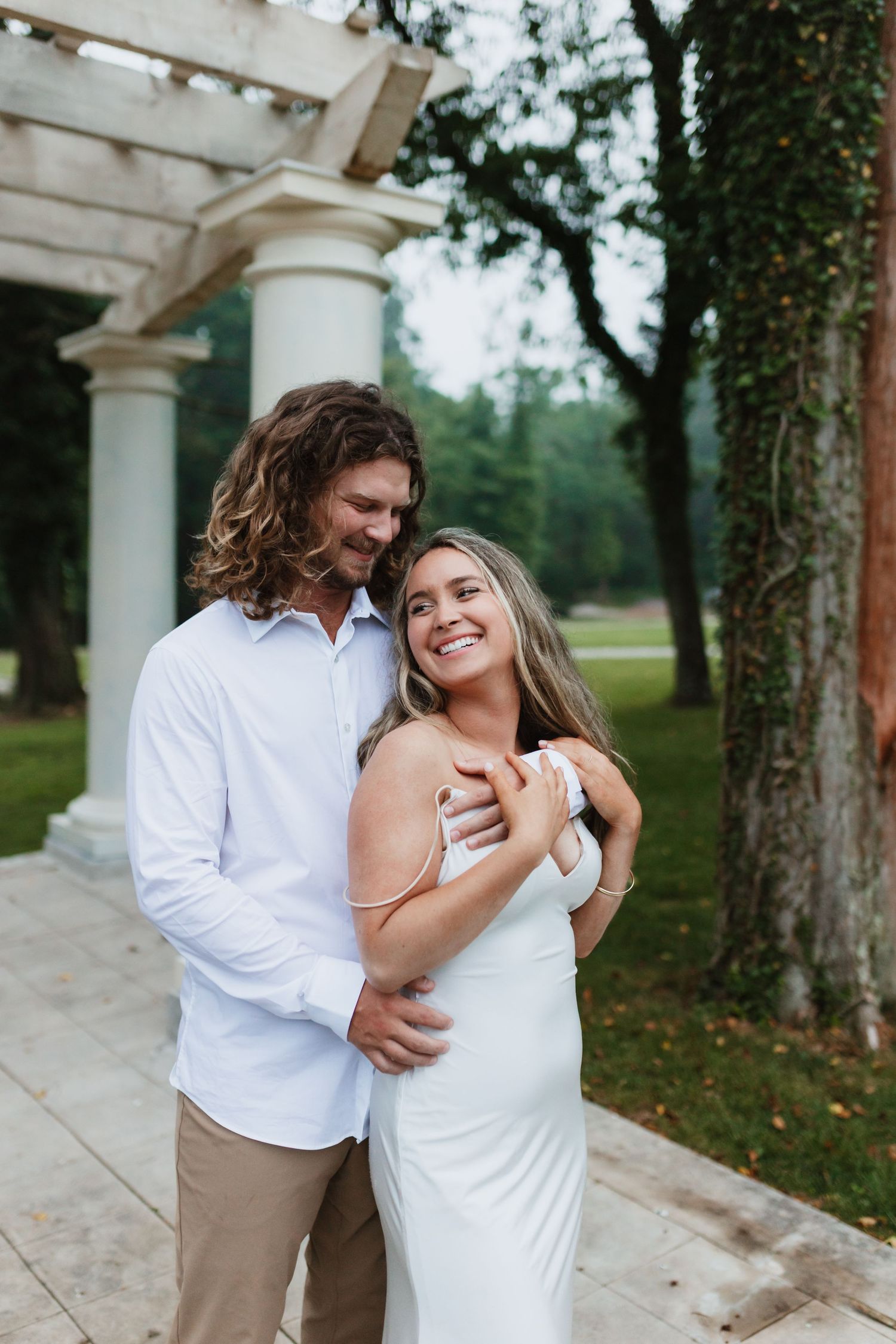 Happy couple embracing near white columns in a garden, woman in white dress smiling up at her partner.