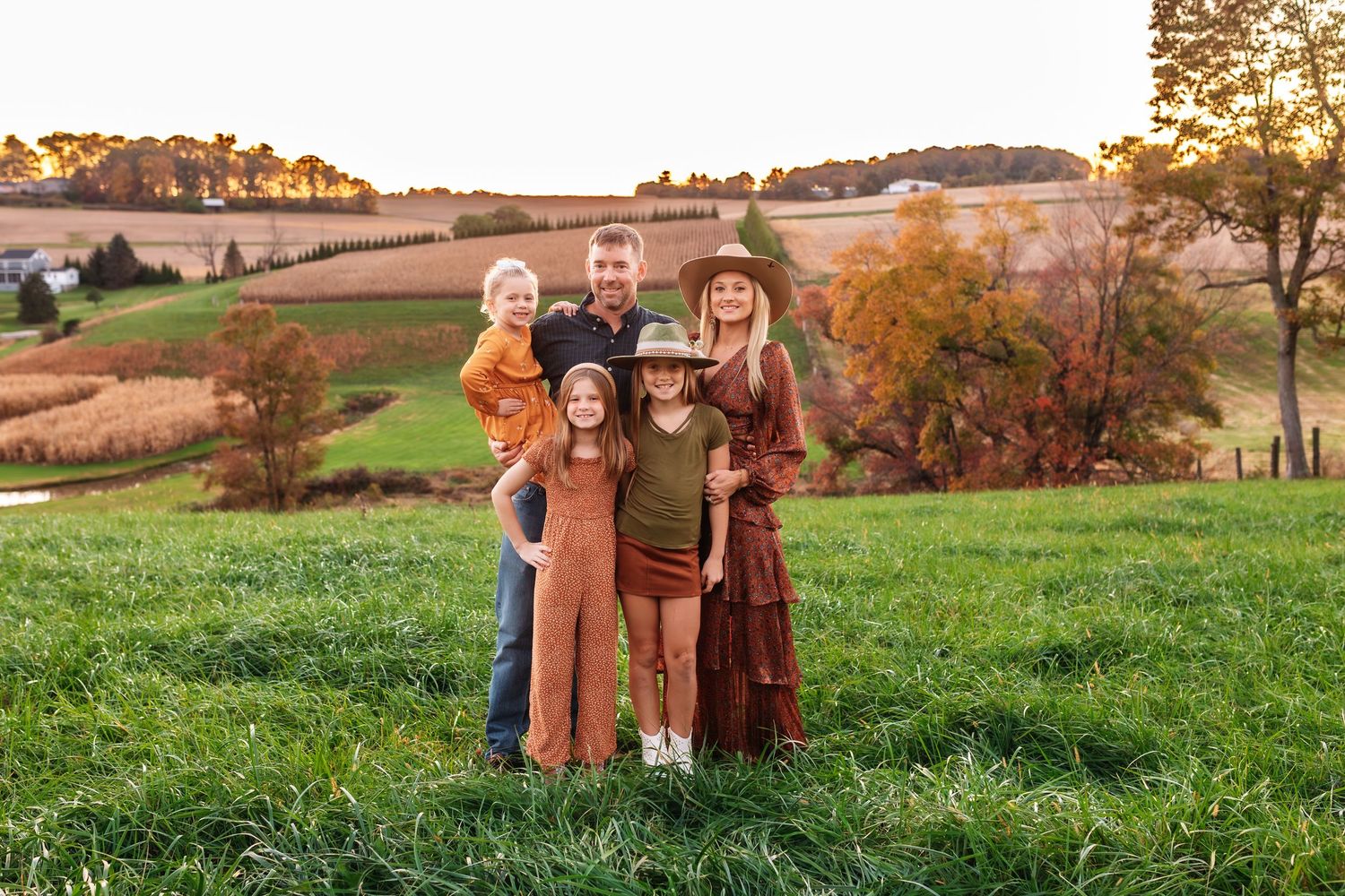 Family of five posing on green hillside with fall foliage background.