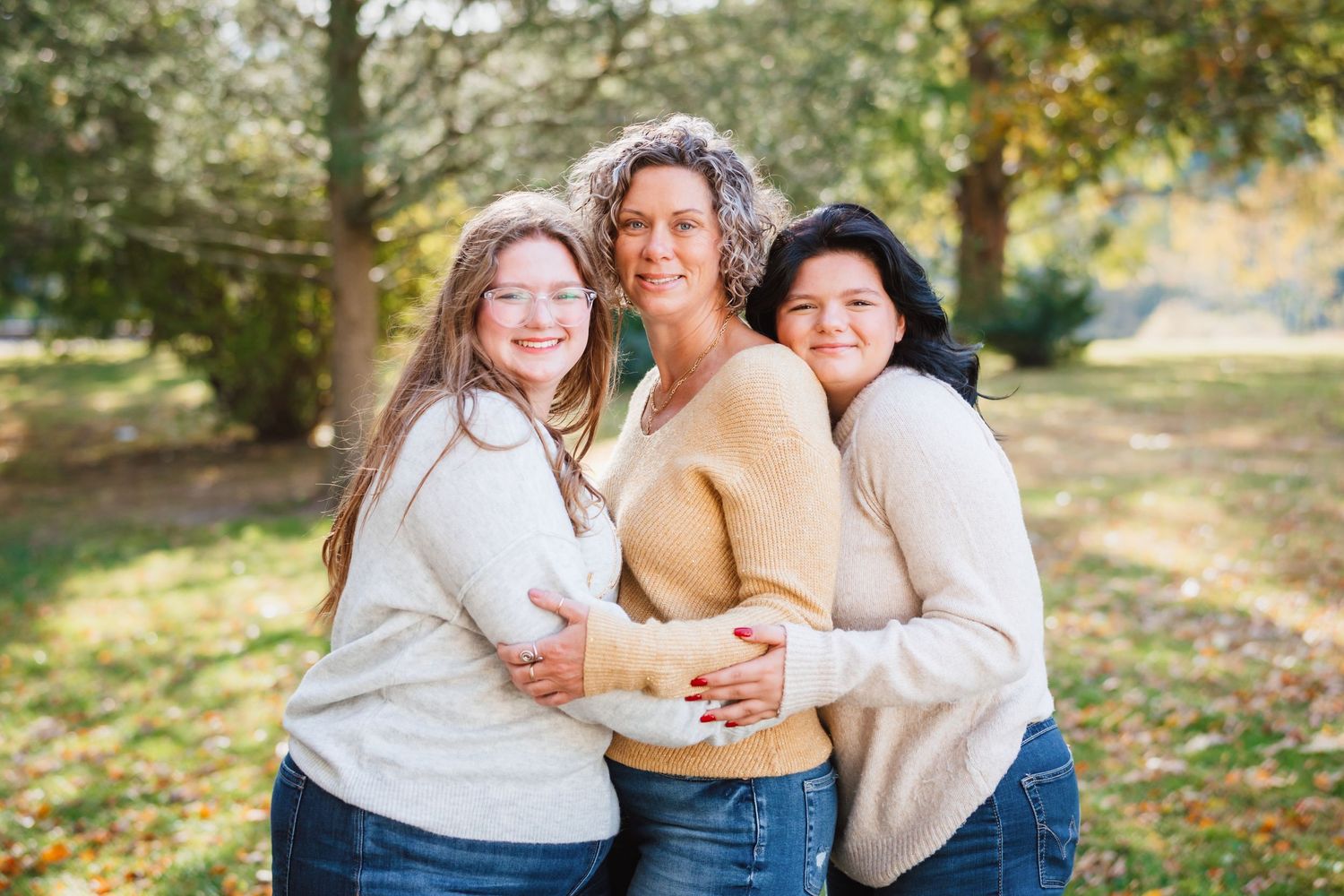 Mother and two teenage daughters embrace warmly while smiling outdoors in a sunny park.