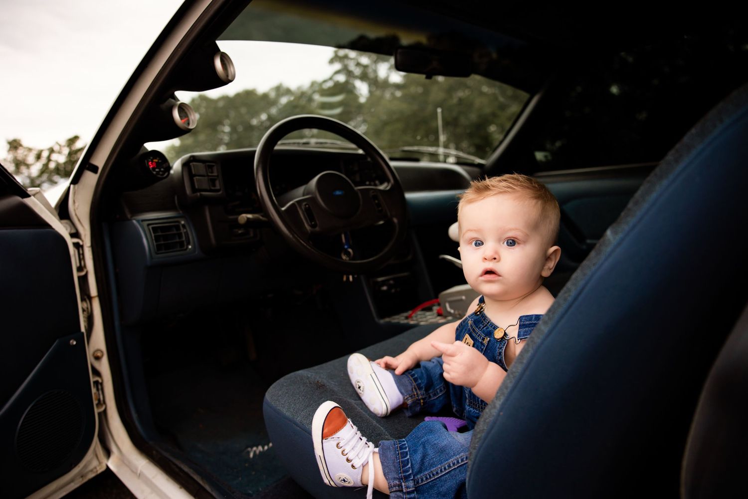 Baby in denim overalls sitting in the front seat of a white car.
