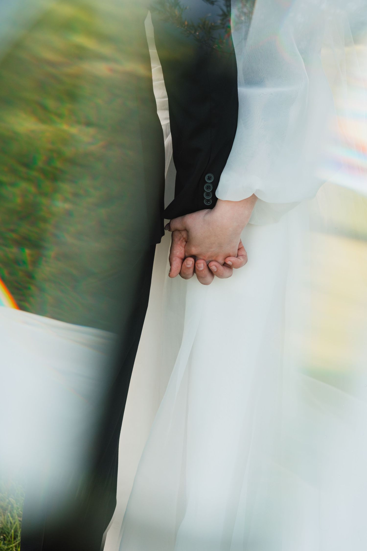 A bride and groom holding hands, showcasing their wedding attire in a softly blurred outdoor setting.