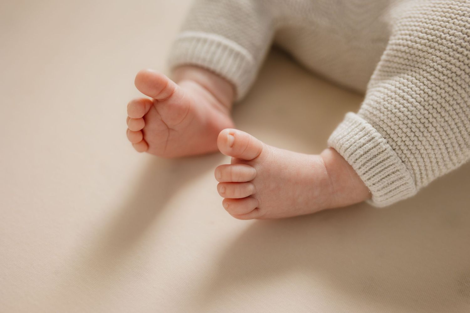 Close-up of a newborn baby's tiny feet and hands in a soft white knit outfit on a beige background.