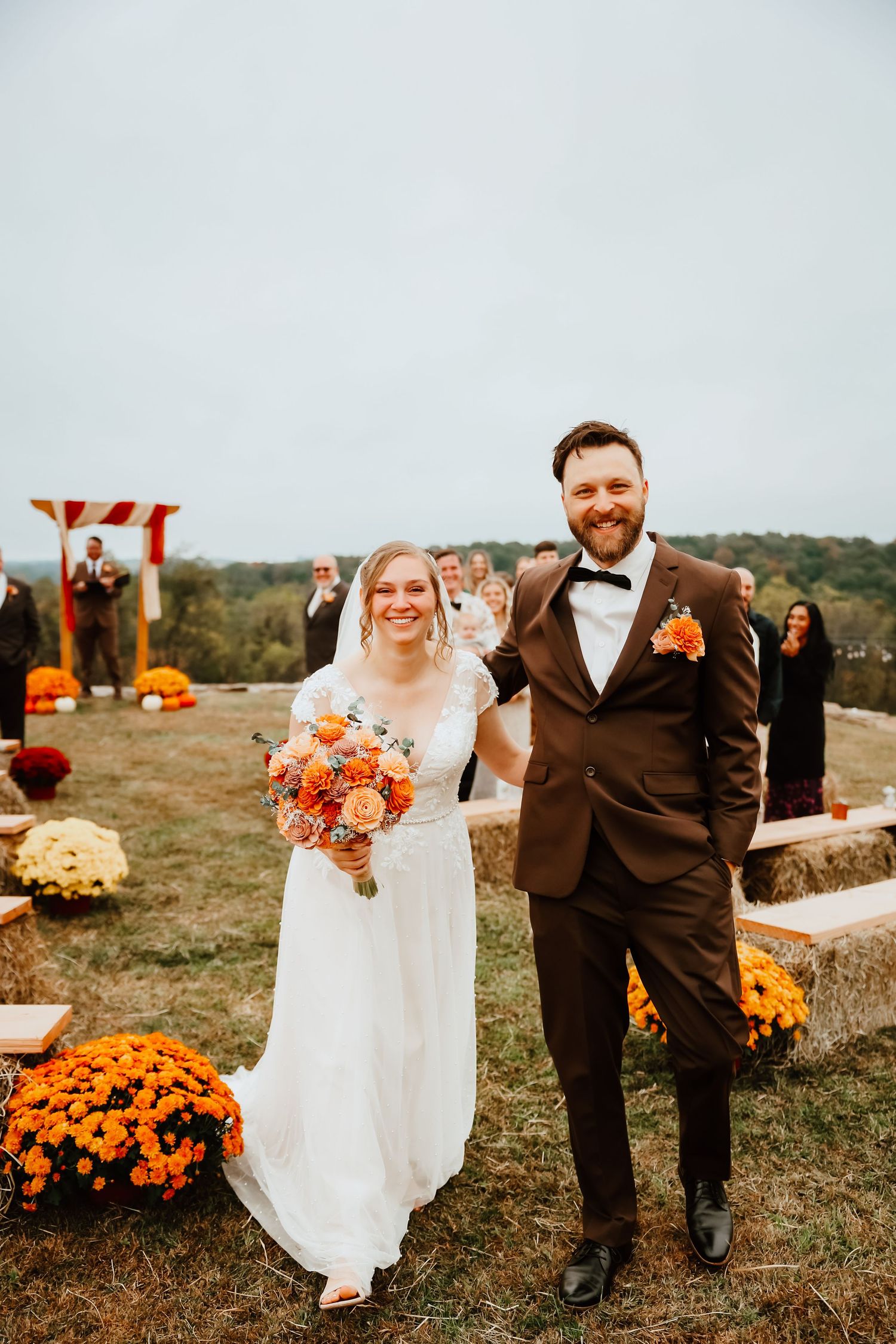 Smiling bride and groom walk down aisle at outdoor fall wedding with orange mum decorations.