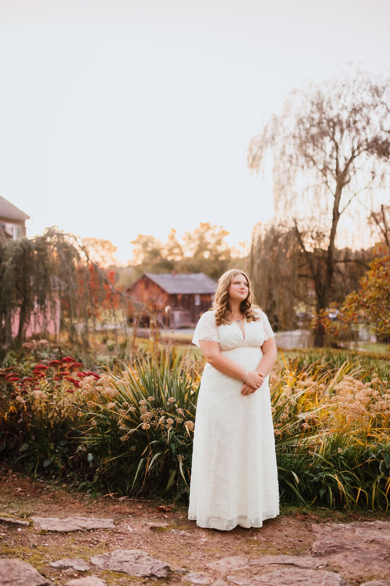 Plus-size bride in white lace dress stands in colorful autumn garden at golden hour.