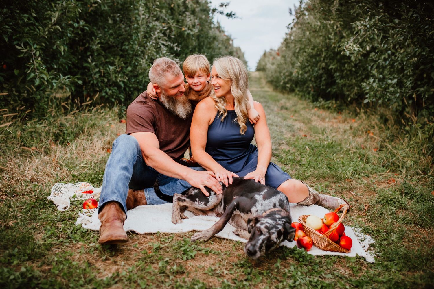 Family of three sitting on a blanket in an orchard with their dog, enjoying a relaxed autumn day.