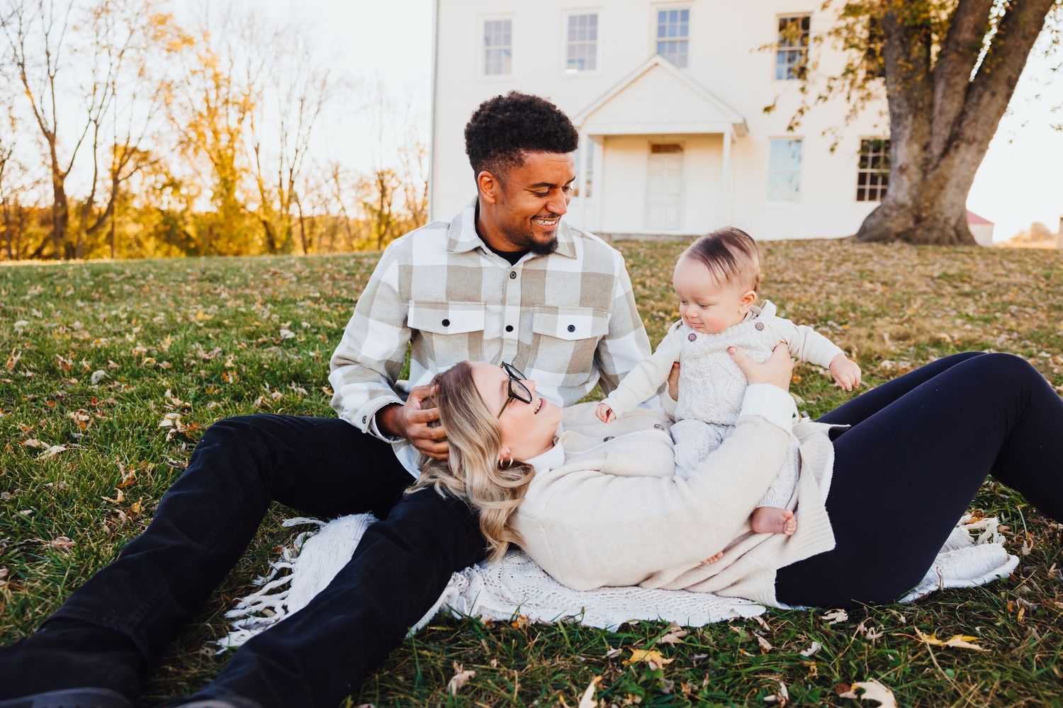 Young interracial family relaxing on a blanket outdoors, parents smiling at their baby in warm autumn light.