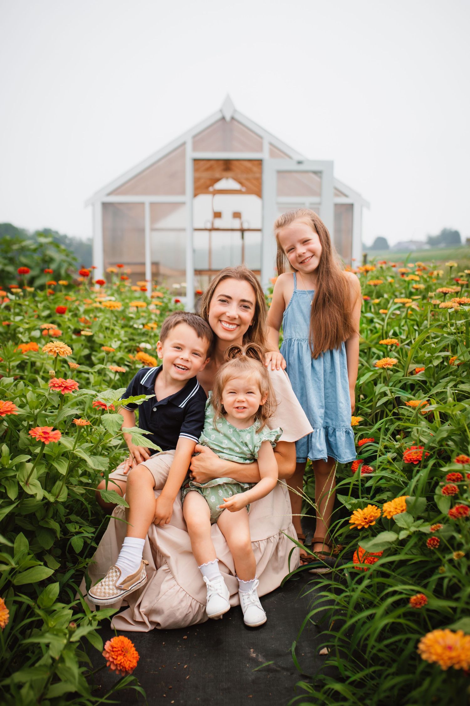 Smiling mother with three children posing in a vibrant zinnia flower field near a greenhouse.