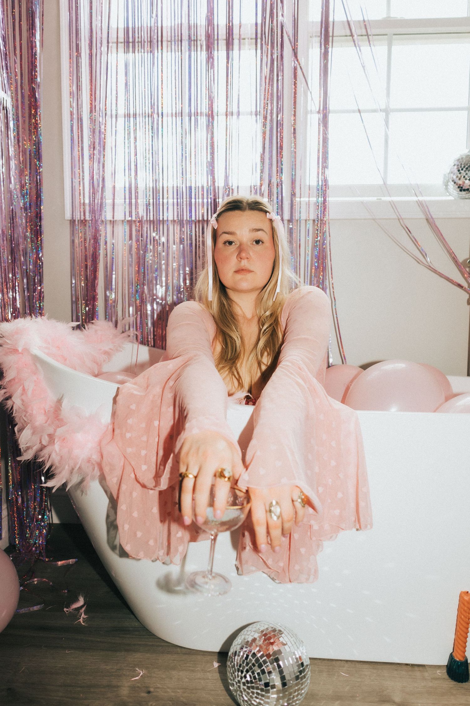 A young blonde woman in pink sitting in a bathtub surrounded by balloons and disco decorations.