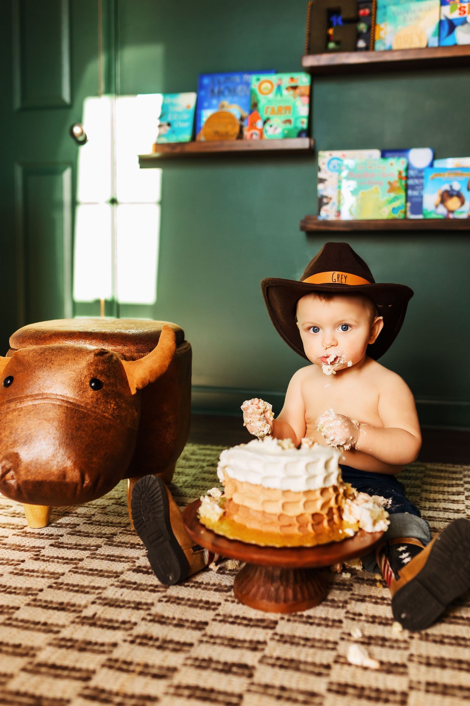 Baby in cowboy hat smashing a layered birthday cake next to a toy bull.