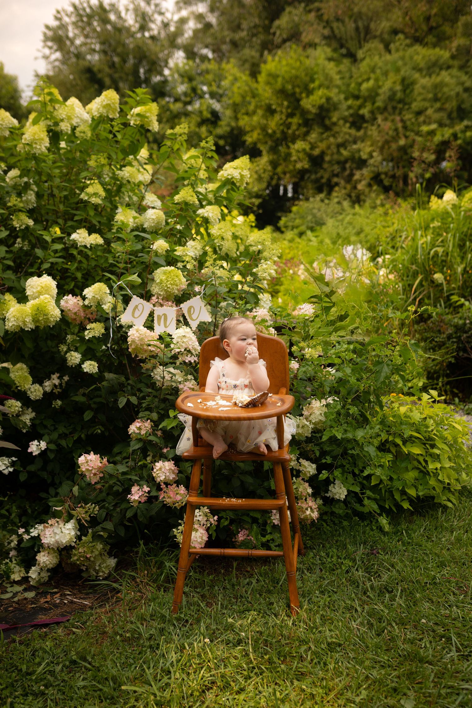 Baby girl in high chair celebrating first birthday with cake in lush garden.