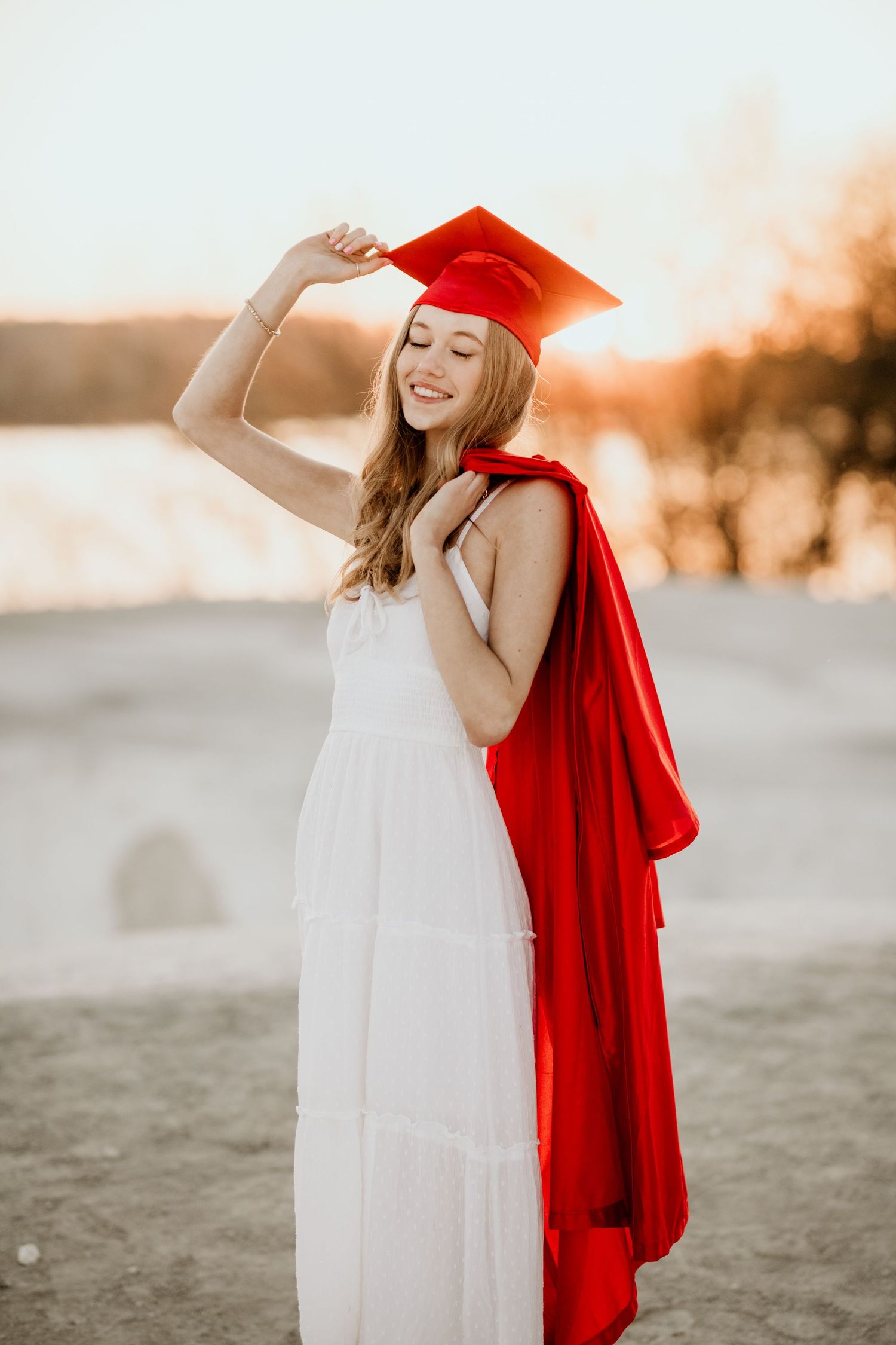 Smiling graduate in red cap and gown over white dress posing outdoors at golden hour.