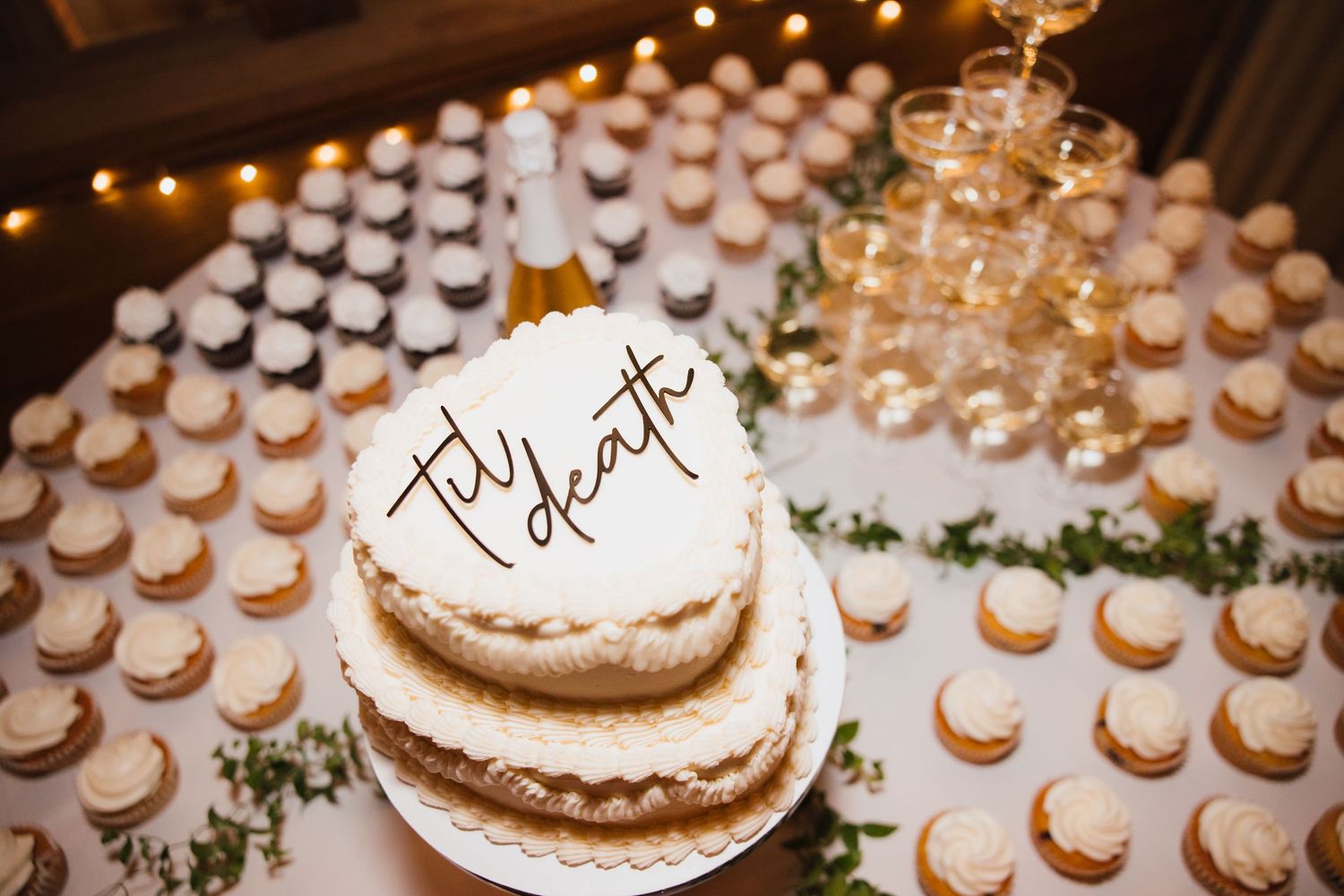 Two-tier wedding cake reading til death surrounded by cupcakes and champagne tower.