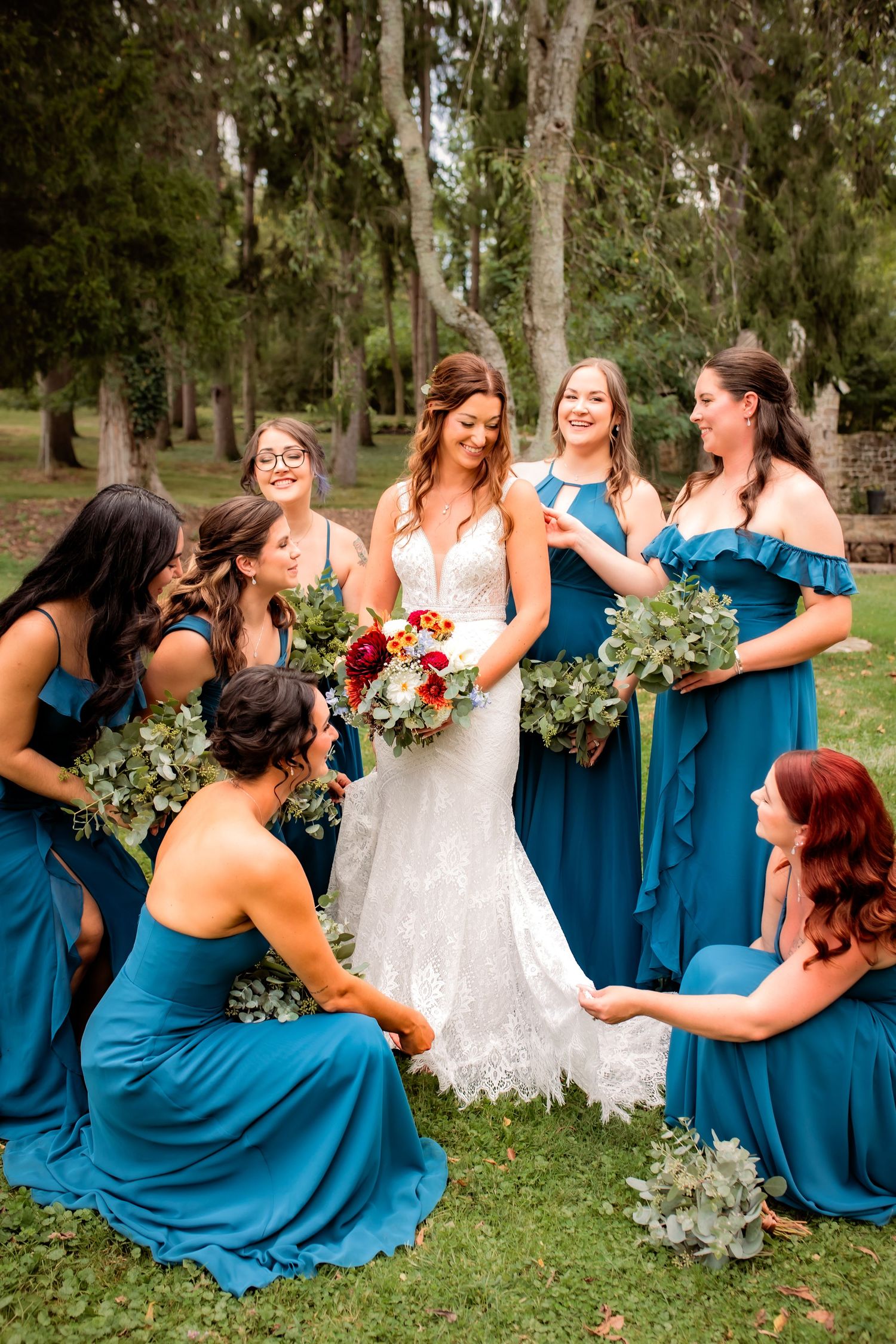 Bride in lace gown surrounded by bridesmaids in teal dresses holding bouquets in a garden.