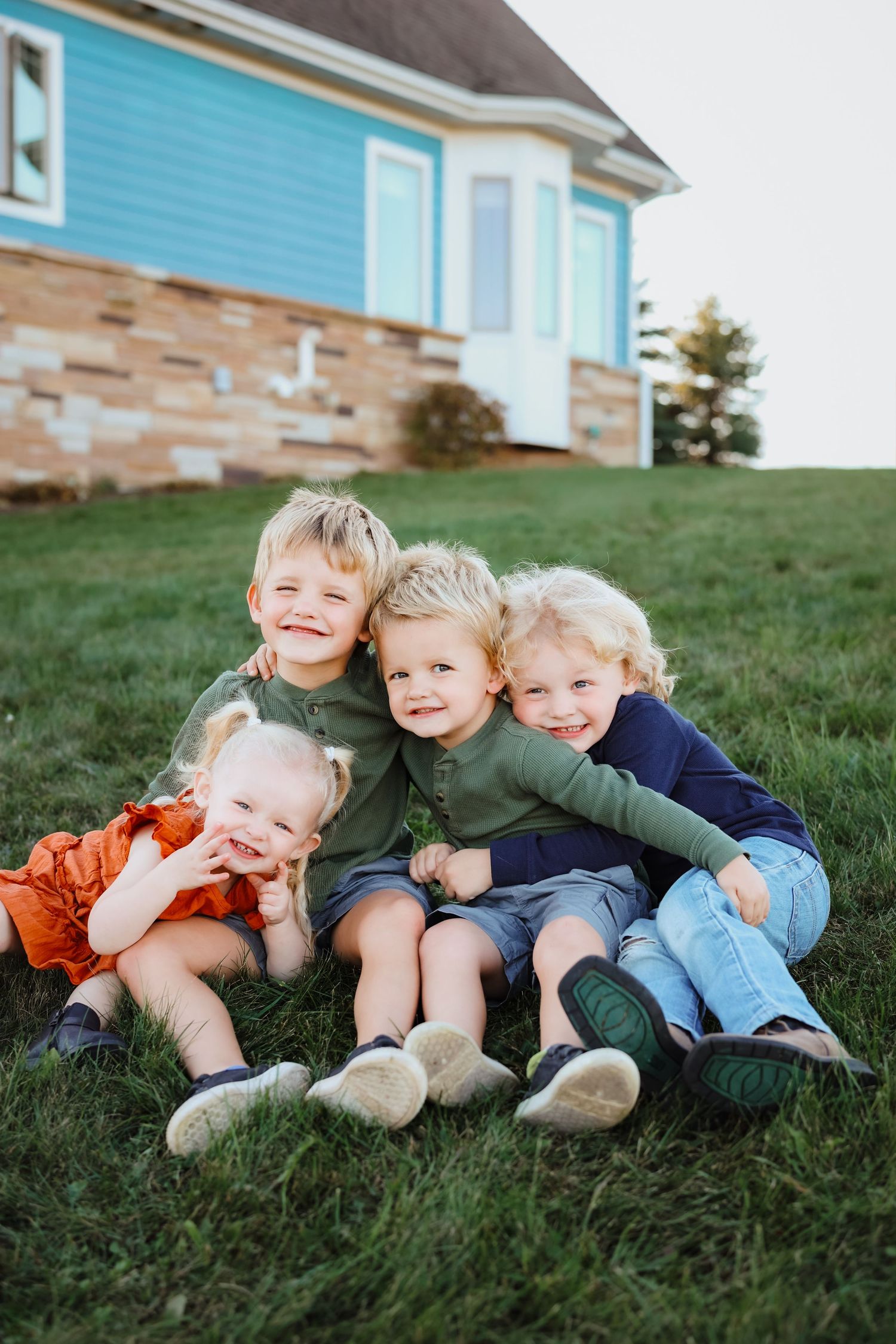 Four young blonde siblings sit together smiling on green grass in front of a blue house.