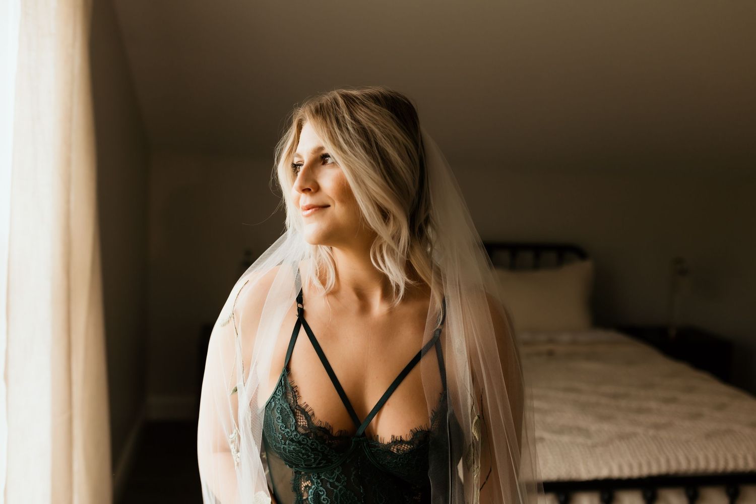 Blonde woman in green lace lingerie and bridal veil gazing out a window in a softly lit bedroom.