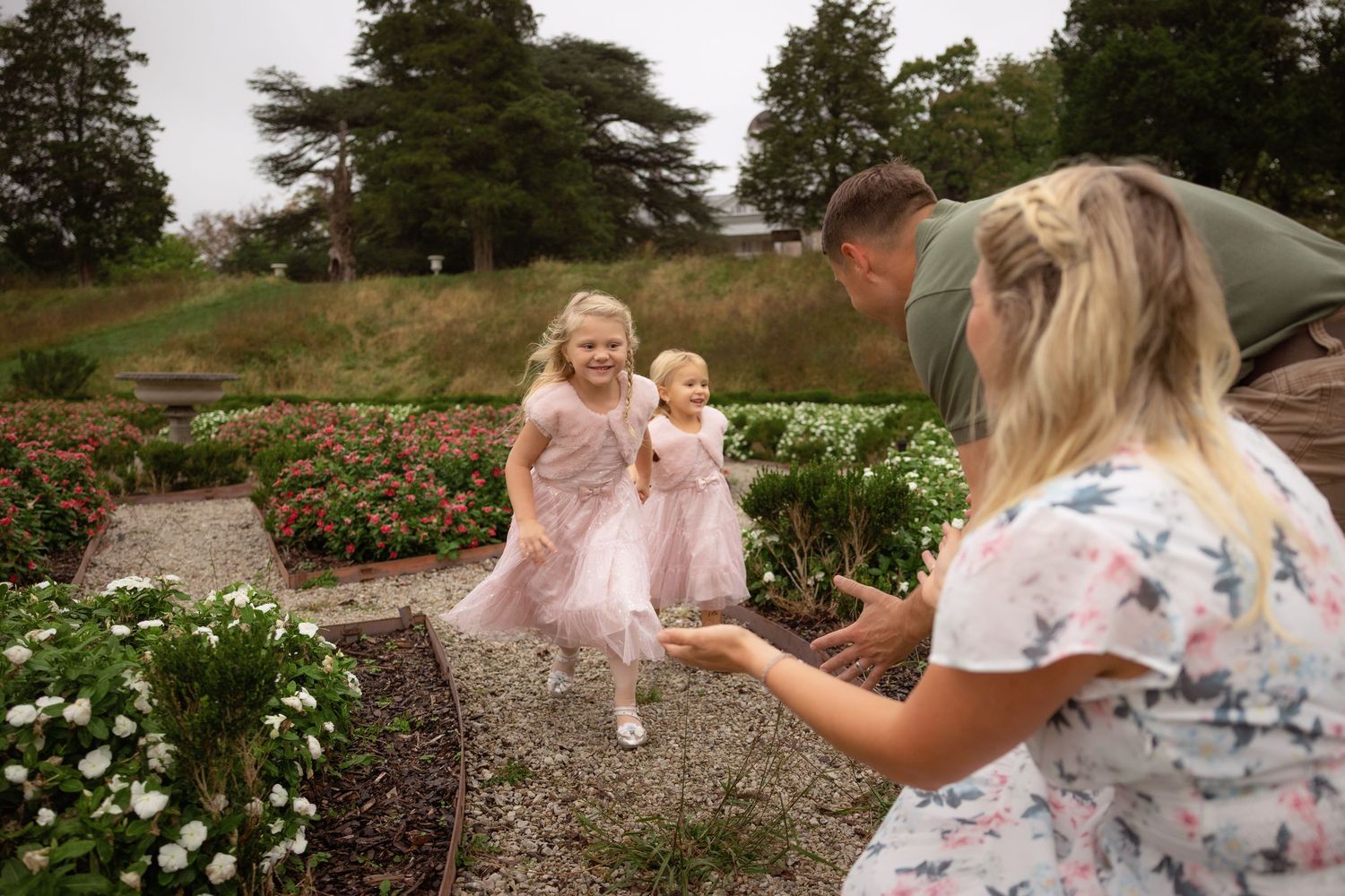 Two little girls in matching pink tulle dresses run along a garden path toward waiting parents, surrounded by flowers and trees.