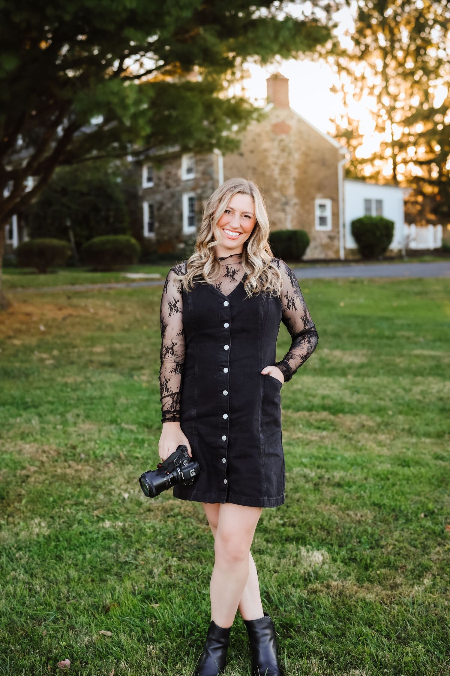 A smiling blonde woman in a black lace dress holding a camera on a green lawn at golden hour.