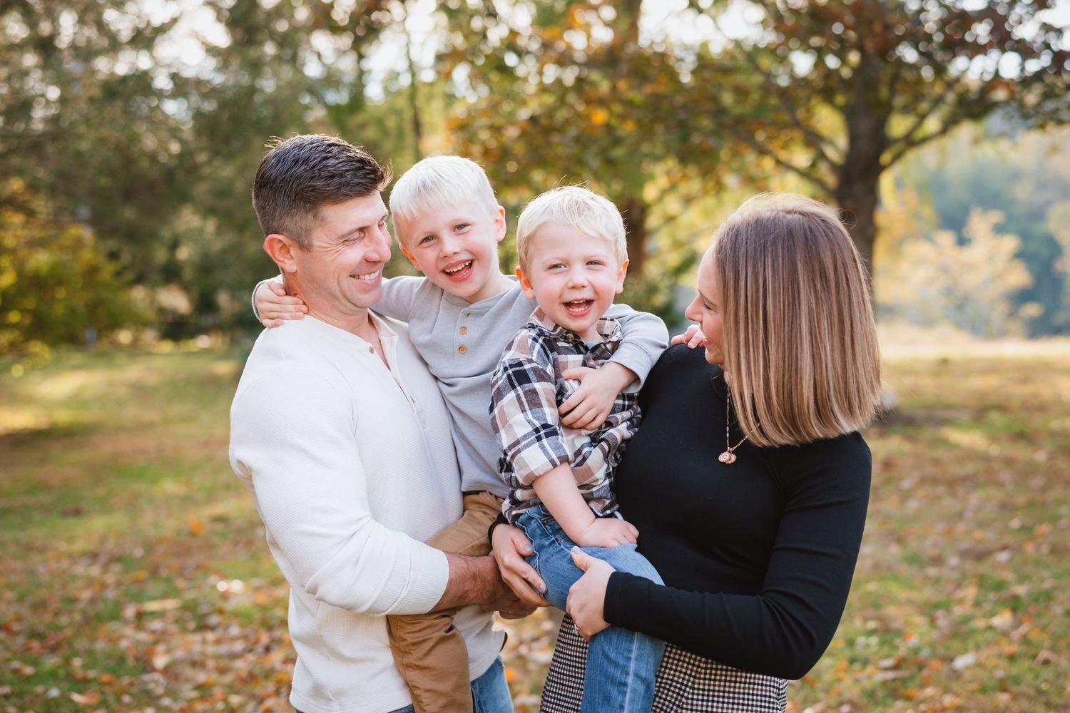 Smiling parents holding two laughing young boys in an autumn park with colorful foliage.