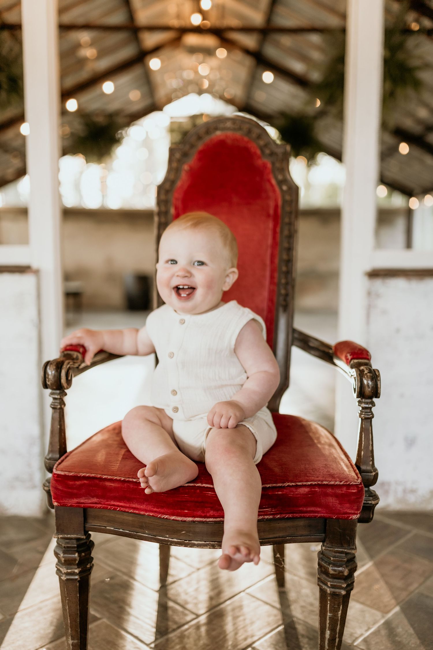 Laughing baby in white romper sitting on ornate red velvet throne chair with festive string lights.
