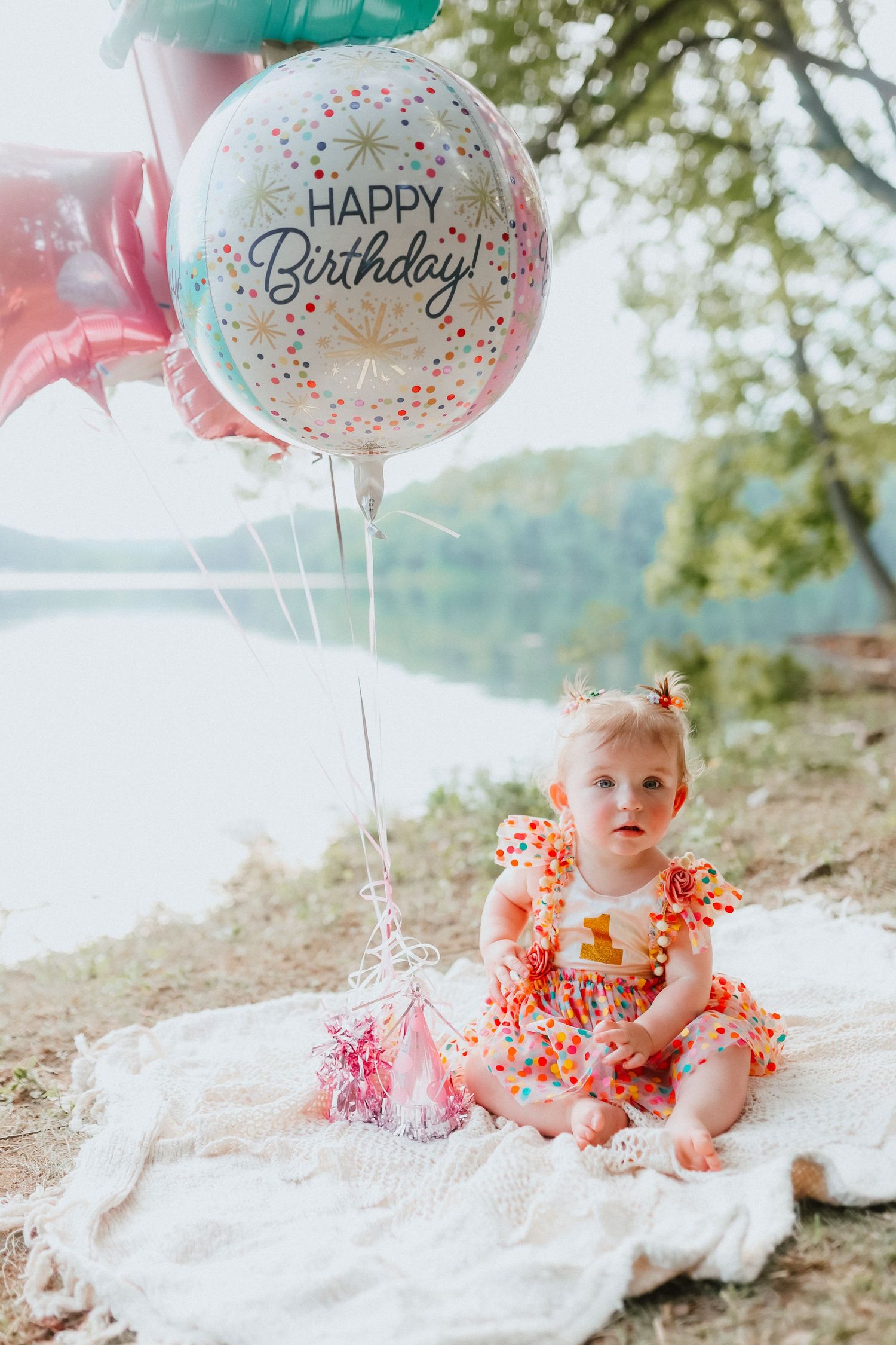 Baby girl in colorful dress sitting outdoors with Happy Birthday balloons by a lake.