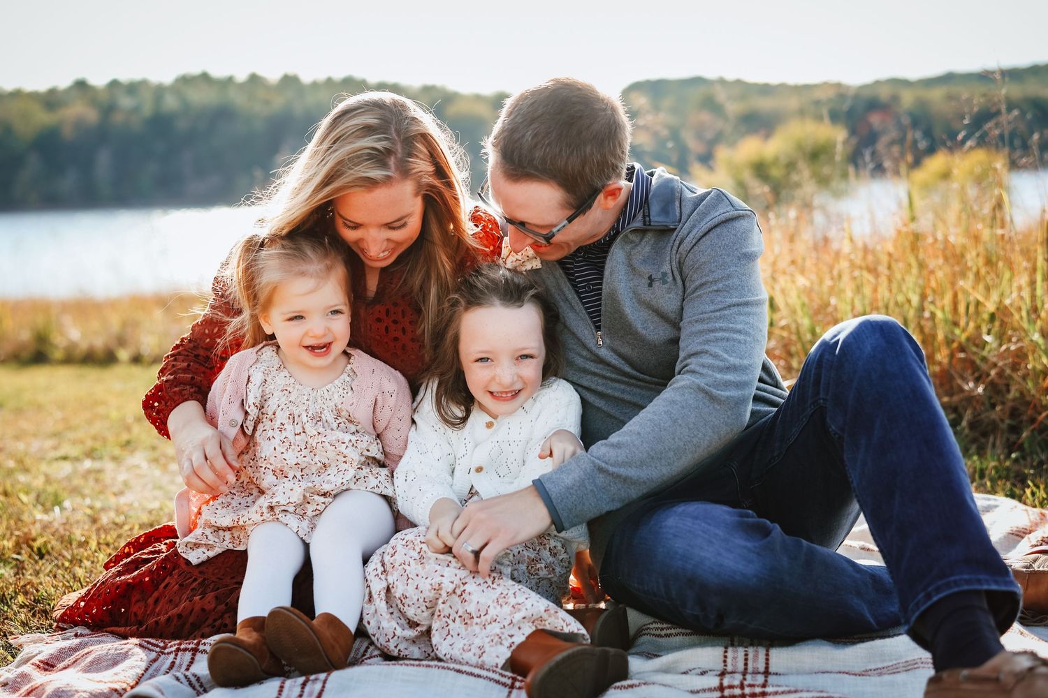 Happy family of four with two toddler girls laughing on a blanket by a lake.