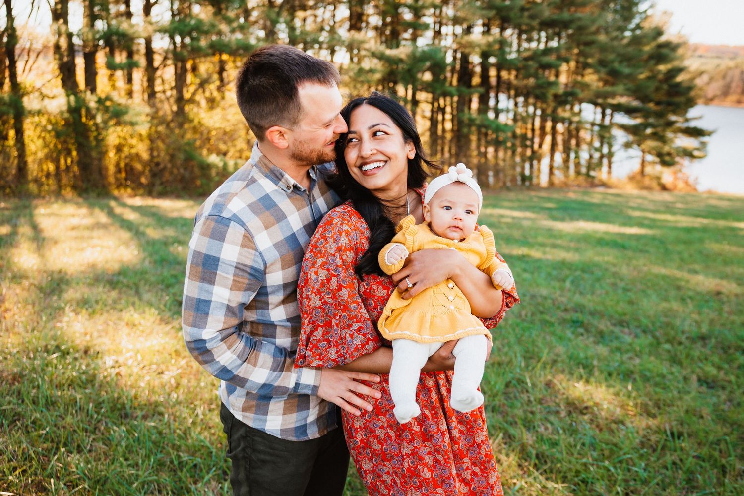 Happy couple holding their baby girl outdoors near a lake, surrounded by colorful autumn trees at sunset.