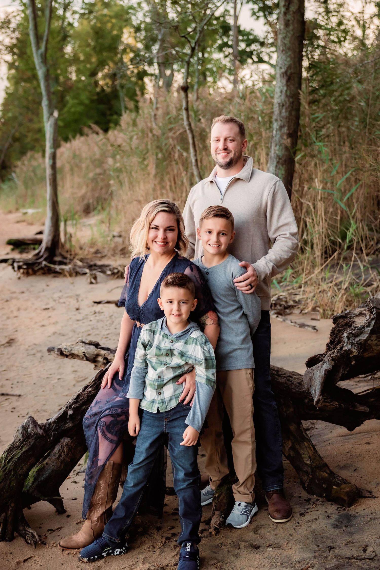Family of four posing together on a fallen log near sandy riverbank surrounded by autumn trees.