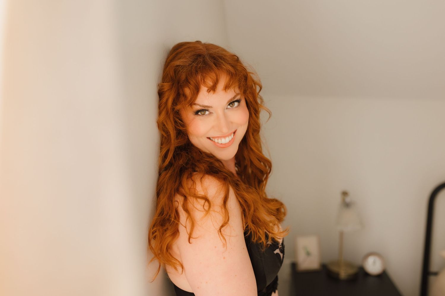 A cheerful redhead woman with curly hair smiles warmly in a bright indoor setting.