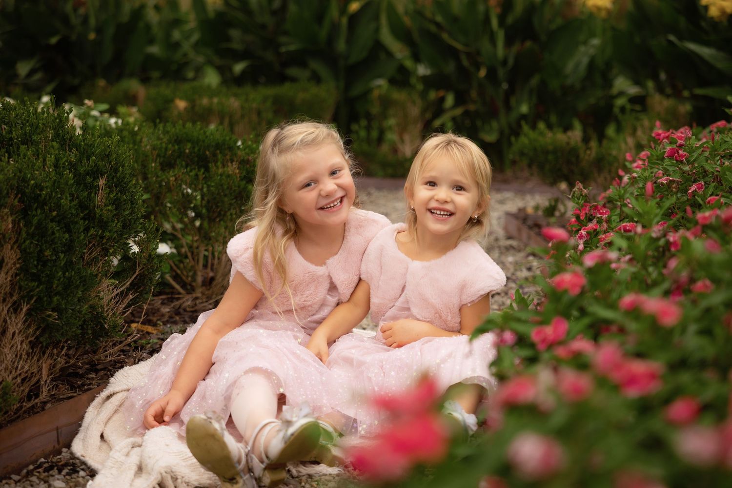 Two smiling young girls in matching pink dresses sit close together on the ground beside blooming pink roses in a lush green garden.