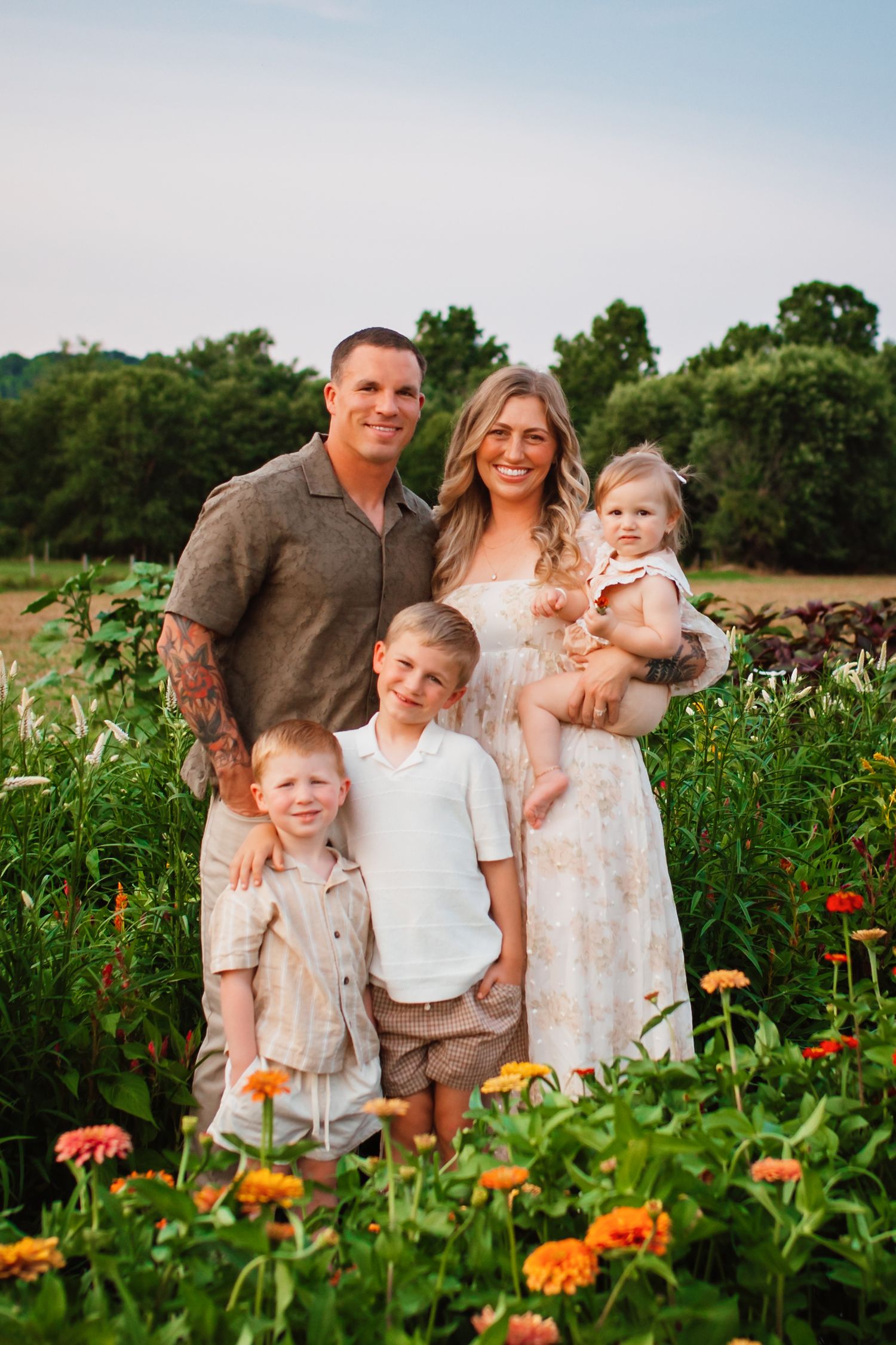 Happy family of five posing in colorful flower field at golden hour, parents with three young children smiling.
