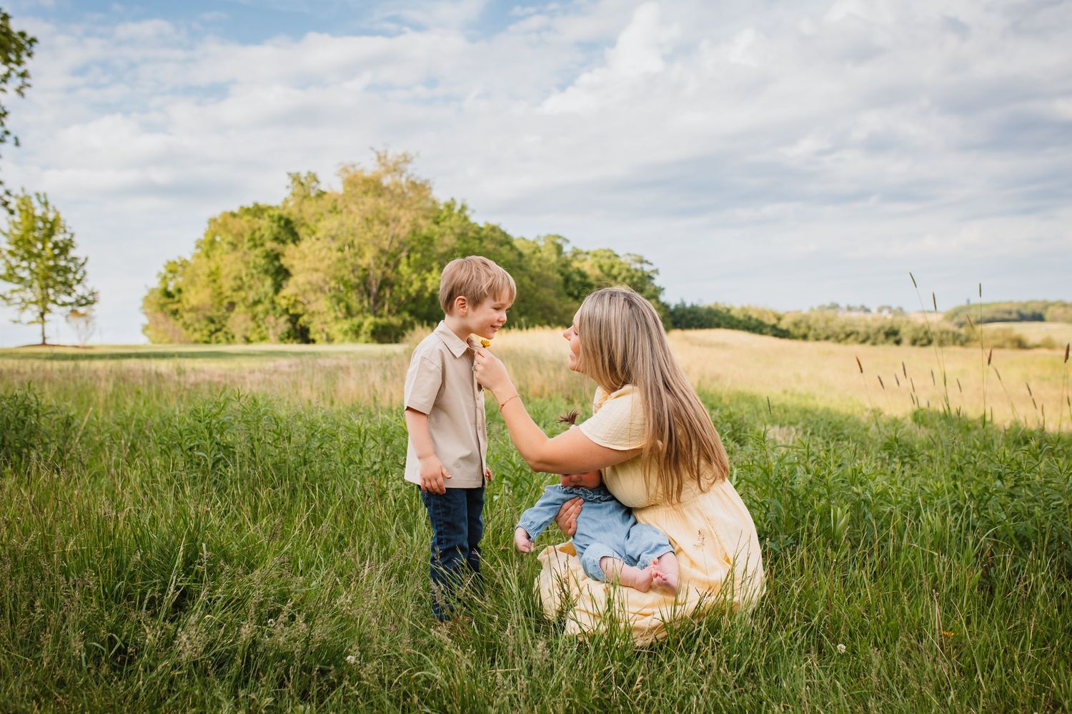 A mother kneels in a green meadow, tenderly interacting with her young son under a cloudy sky.