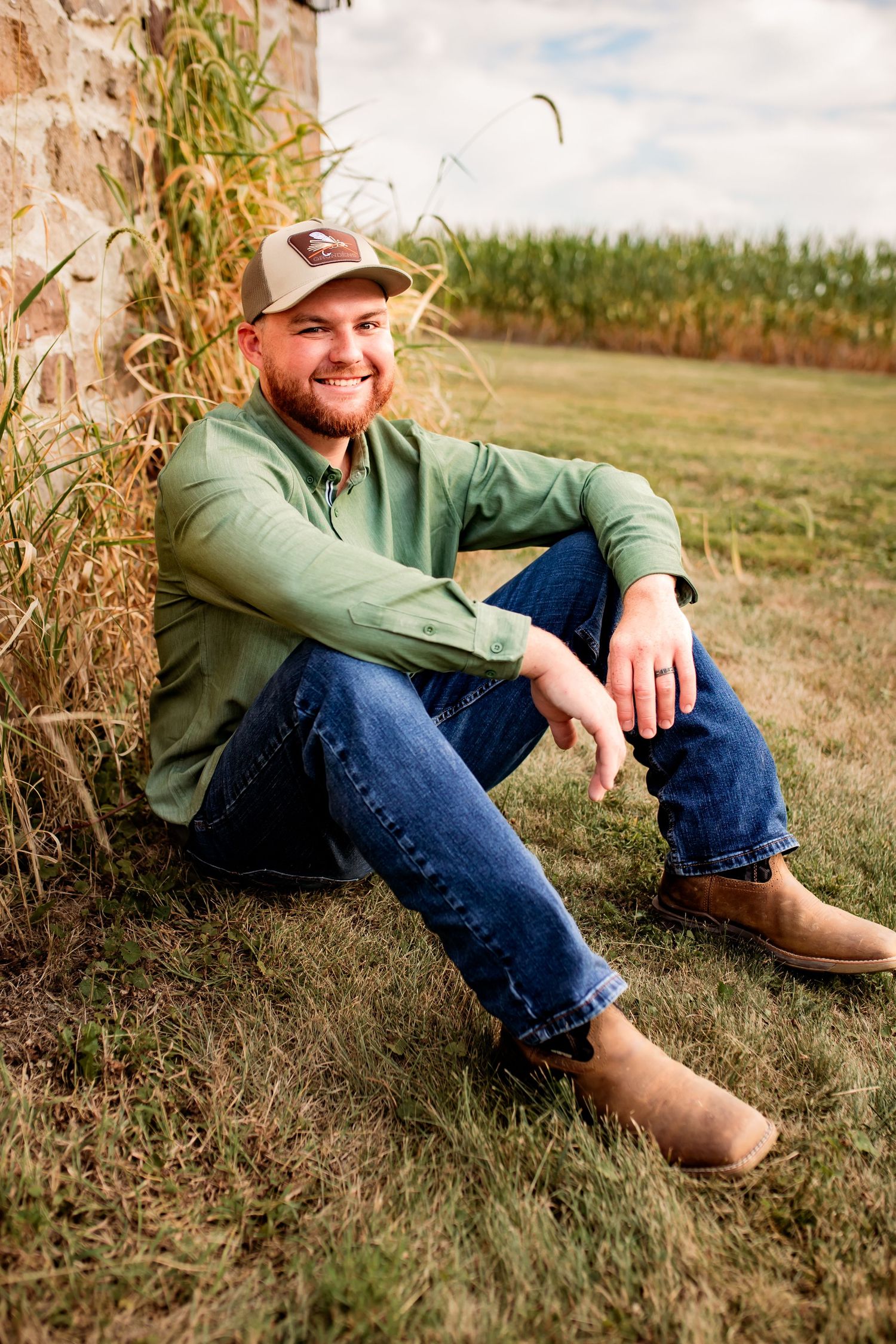 Smiling groom in green shirt and trucker hat sitting against stone wall near cornfield.