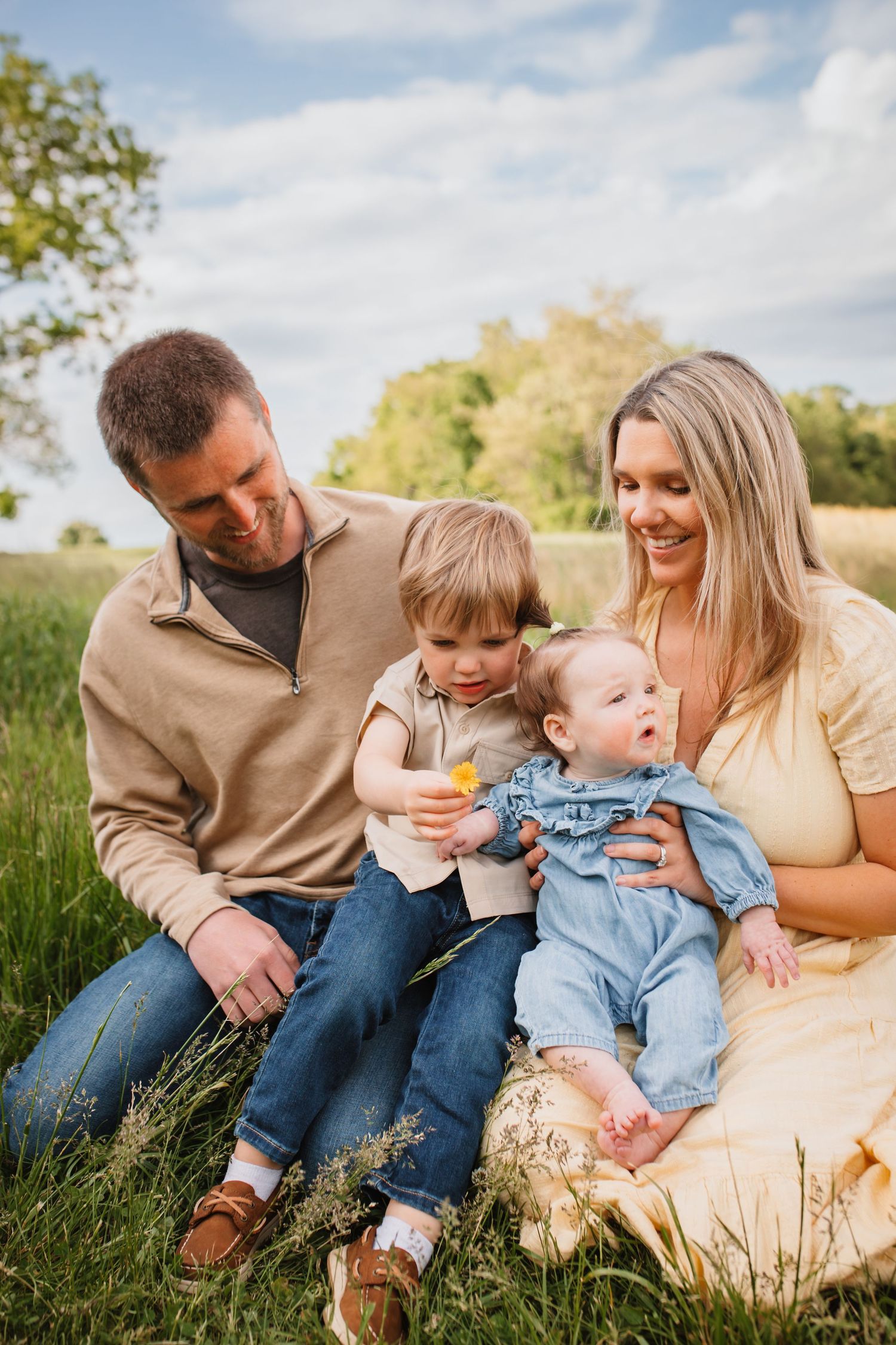 Family of four sitting in a grassy field, parents smiling at their toddler and baby on a sunny day.