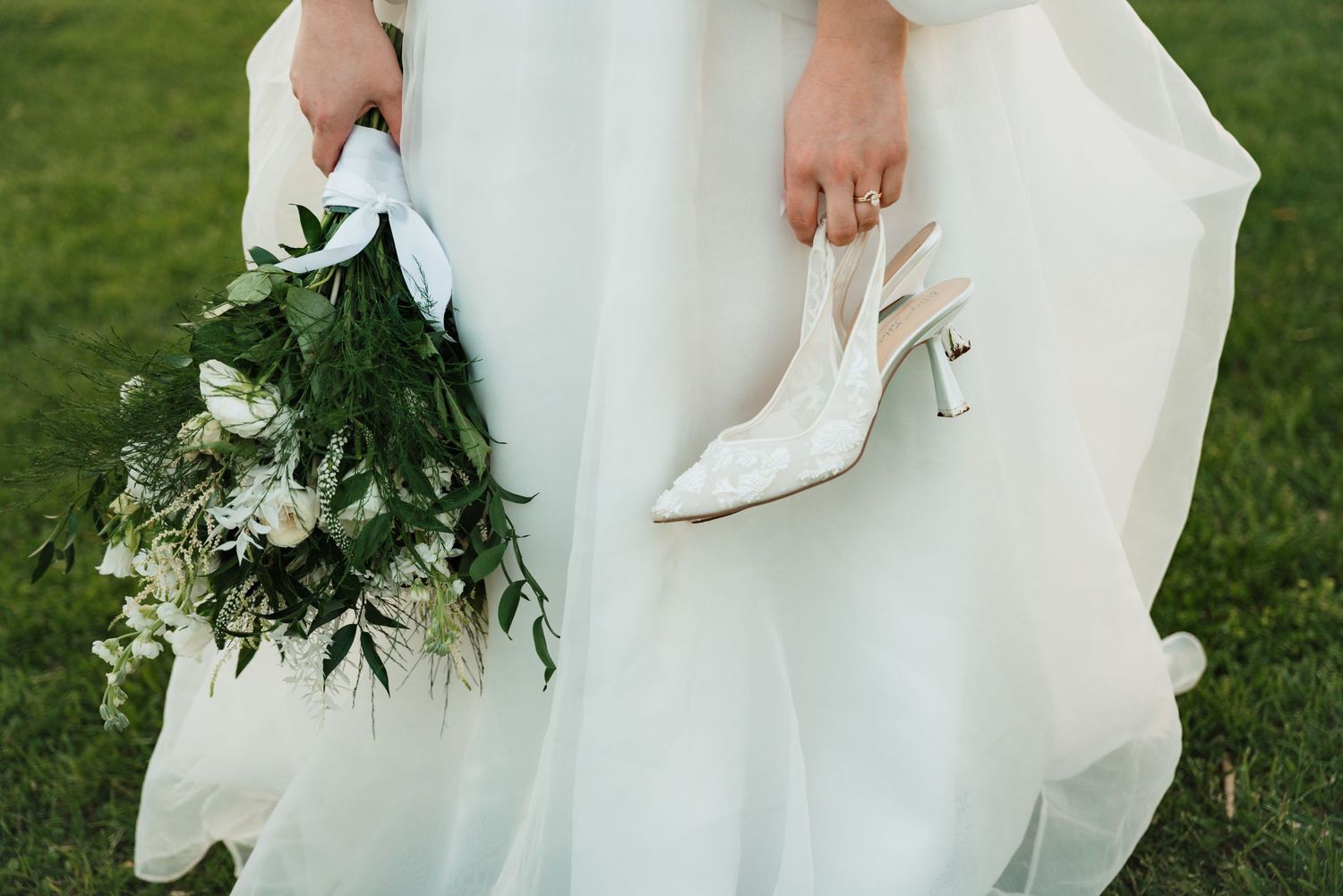 Bride holds white floral bouquet and lace heels against flowing white wedding gown on green grass.