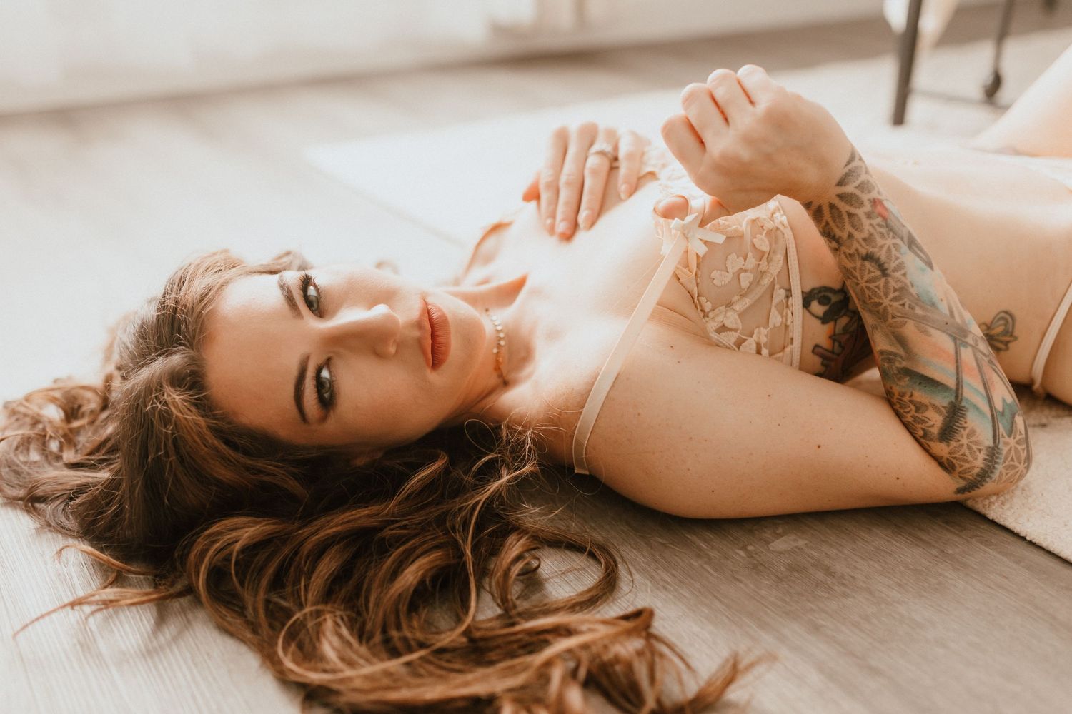Tattooed brunette woman in nude lingerie lying on a hardwood floor with long flowing hair.