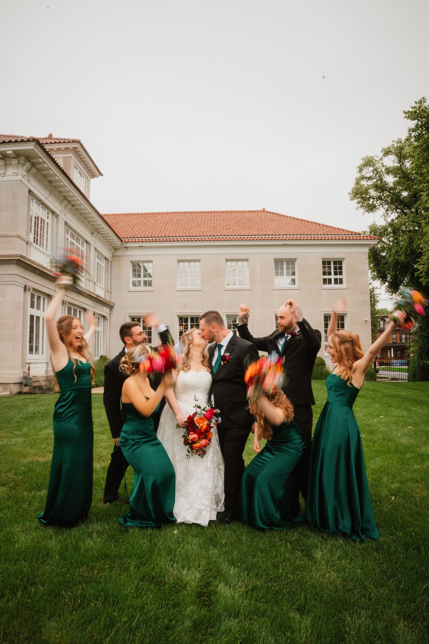 Wedding party in emerald green dresses celebrates with couple kissing on lawn of elegant mansion.
