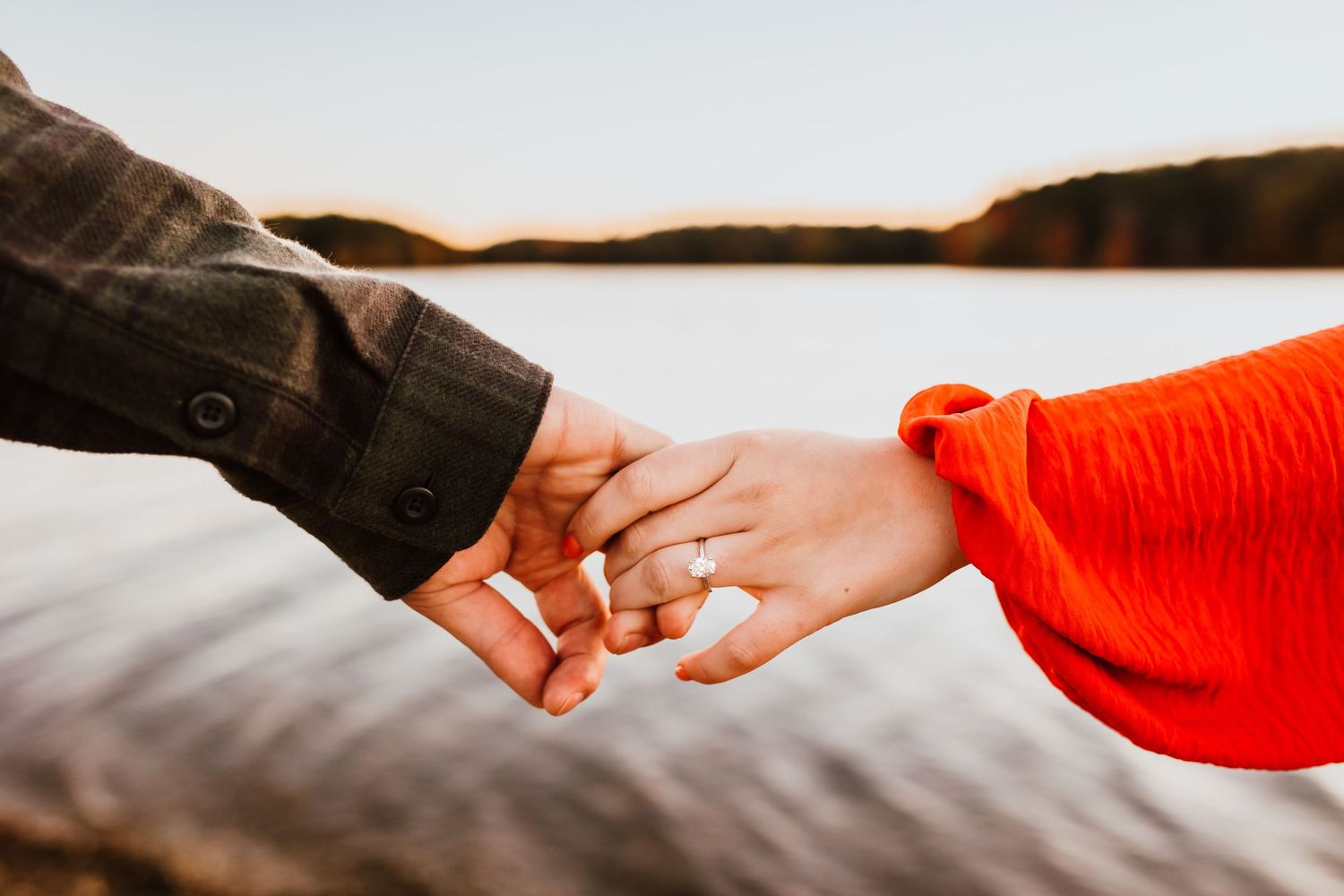 Couple holding hands near a serene lake at sunset, engagement ring visible.