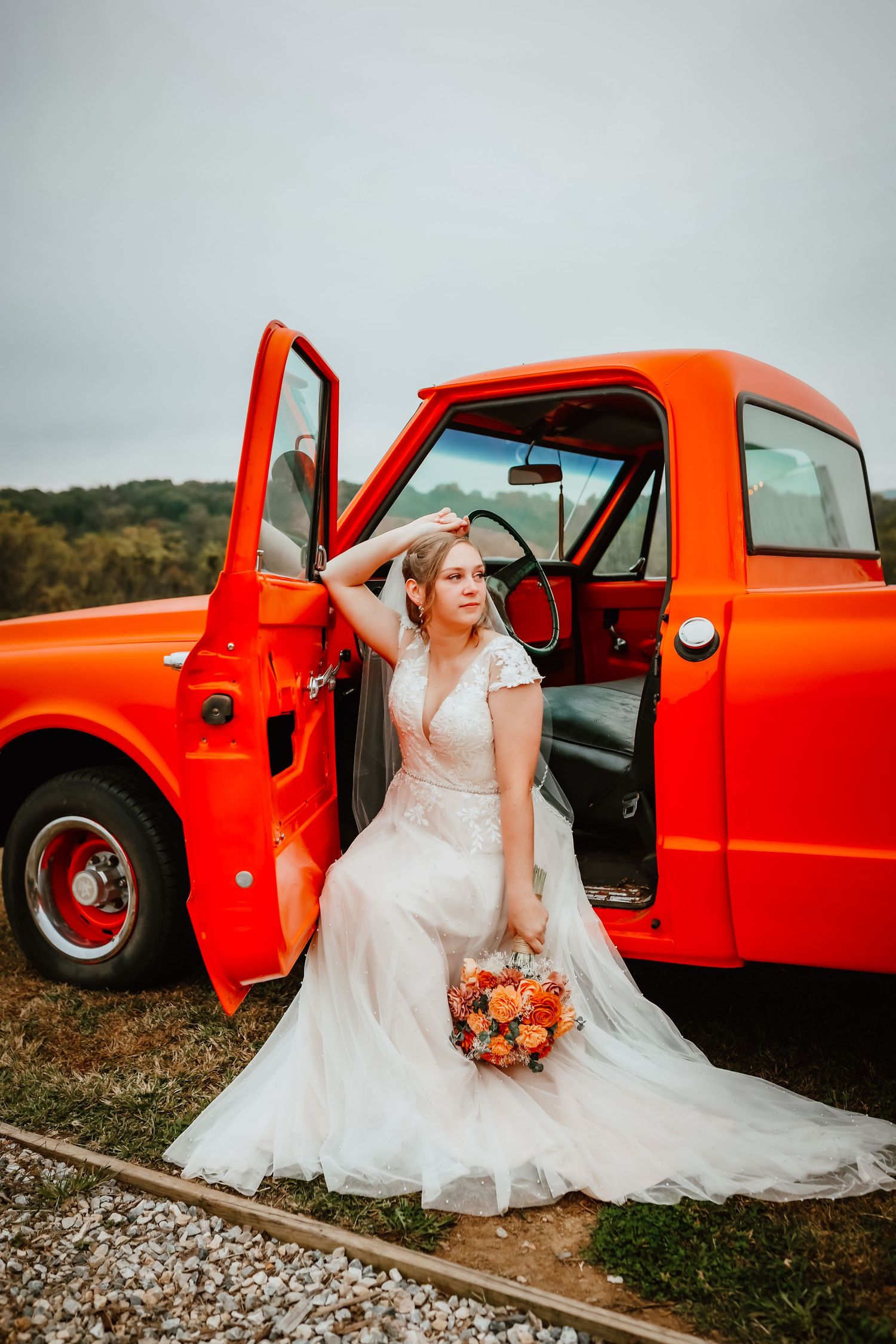 Bride in white lace gown sitting in open door of vintage red truck holding orange fall bouquet.
