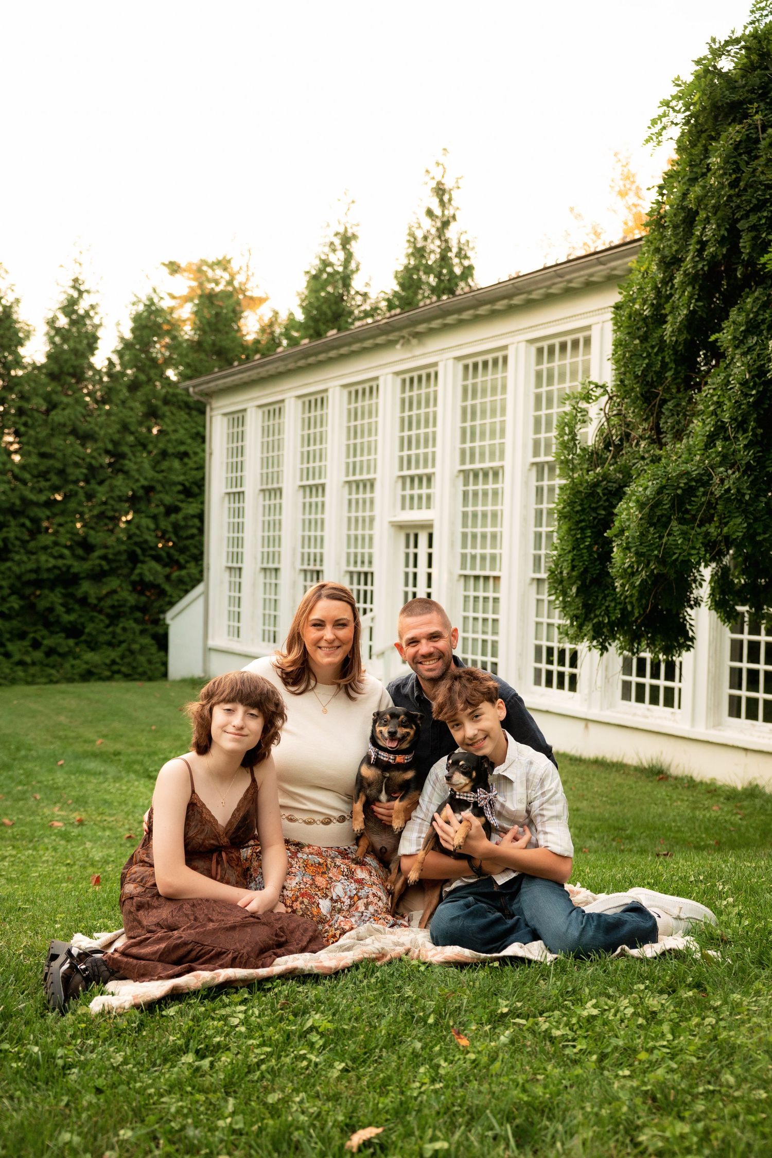 Family of four sitting on a blanket on green grass holding two small dogs in front of a white glass-paned greenhouse surrounded by trees.
