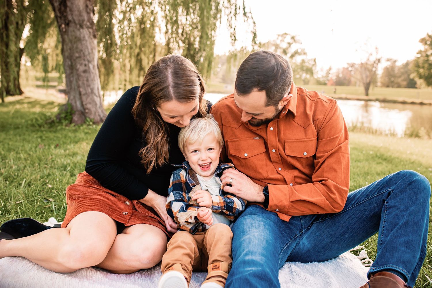 Young family of three sitting on blanket by pond, parents laughing with their toddler son outdoors.