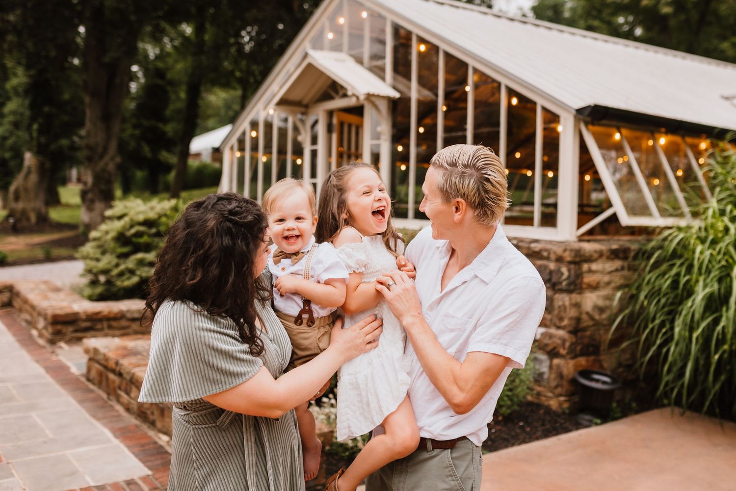 Happy family with two young children laughing together outside a charming greenhouse with string lights.