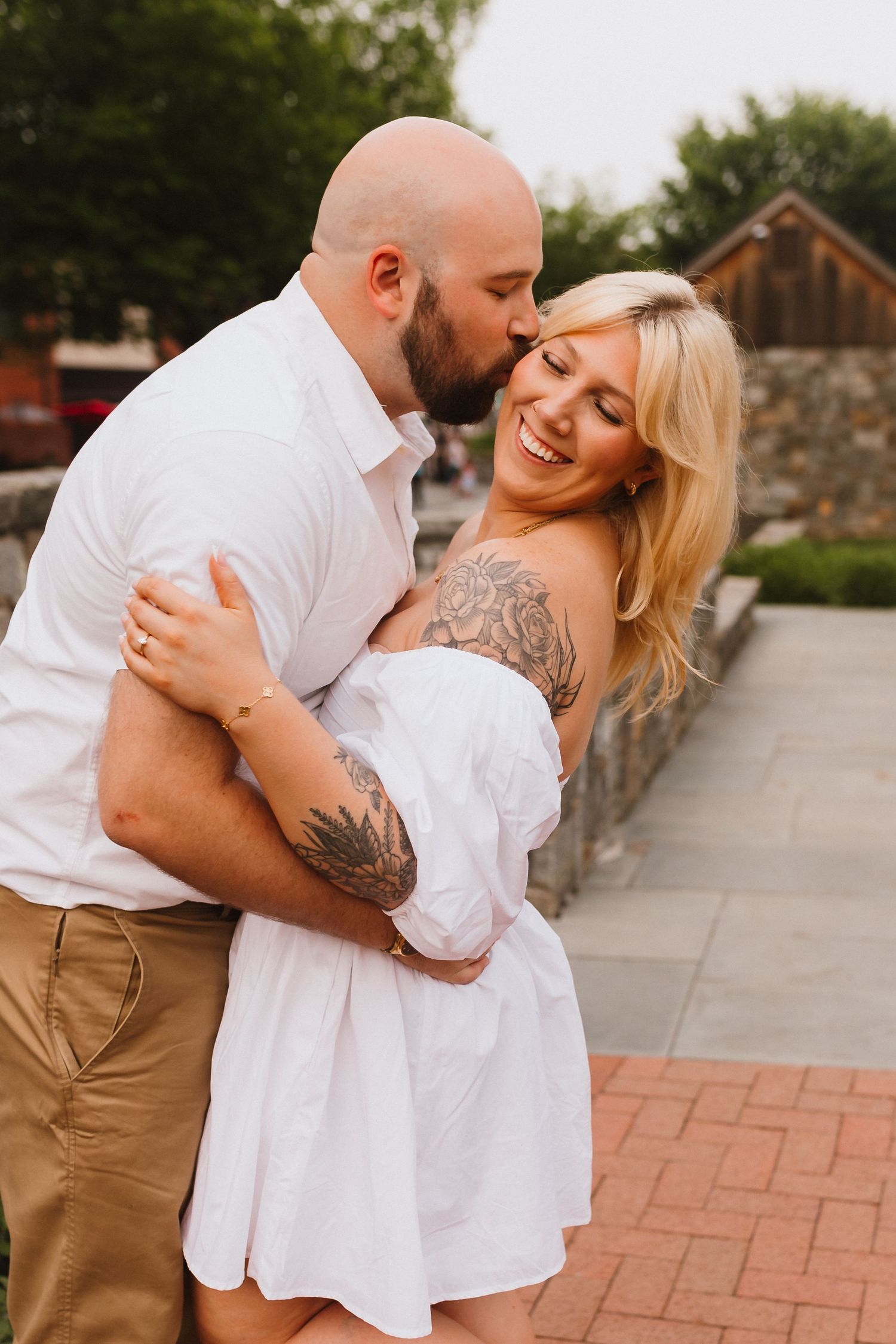 Bald man kissing tattooed blonde woman in white dress outdoors.