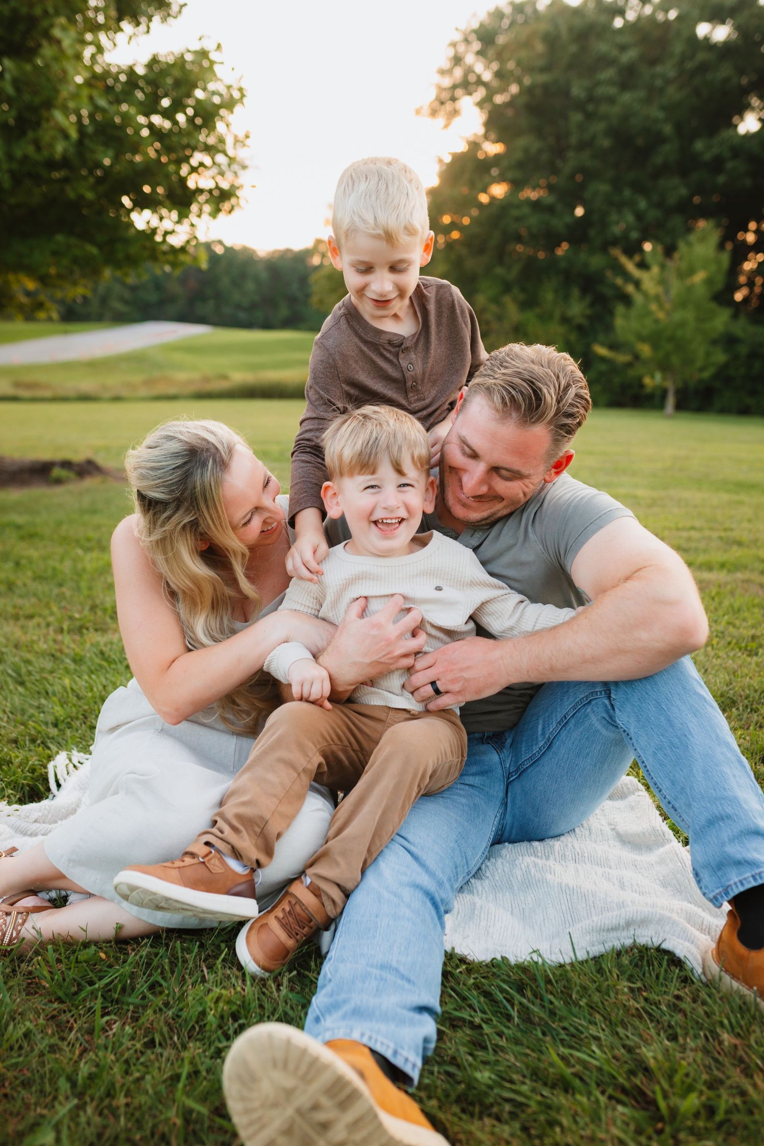 Family of four laughing and cuddling on a blanket in a lush green park at golden hour sunset.