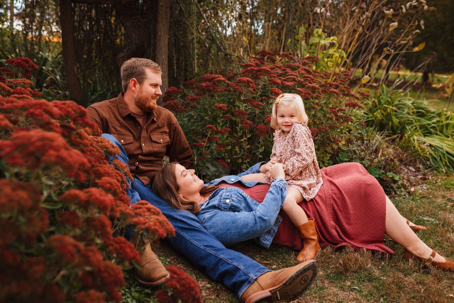 Family of three relaxing outdoors near red flowers, parents smiling at toddler daughter in warm fall setting.