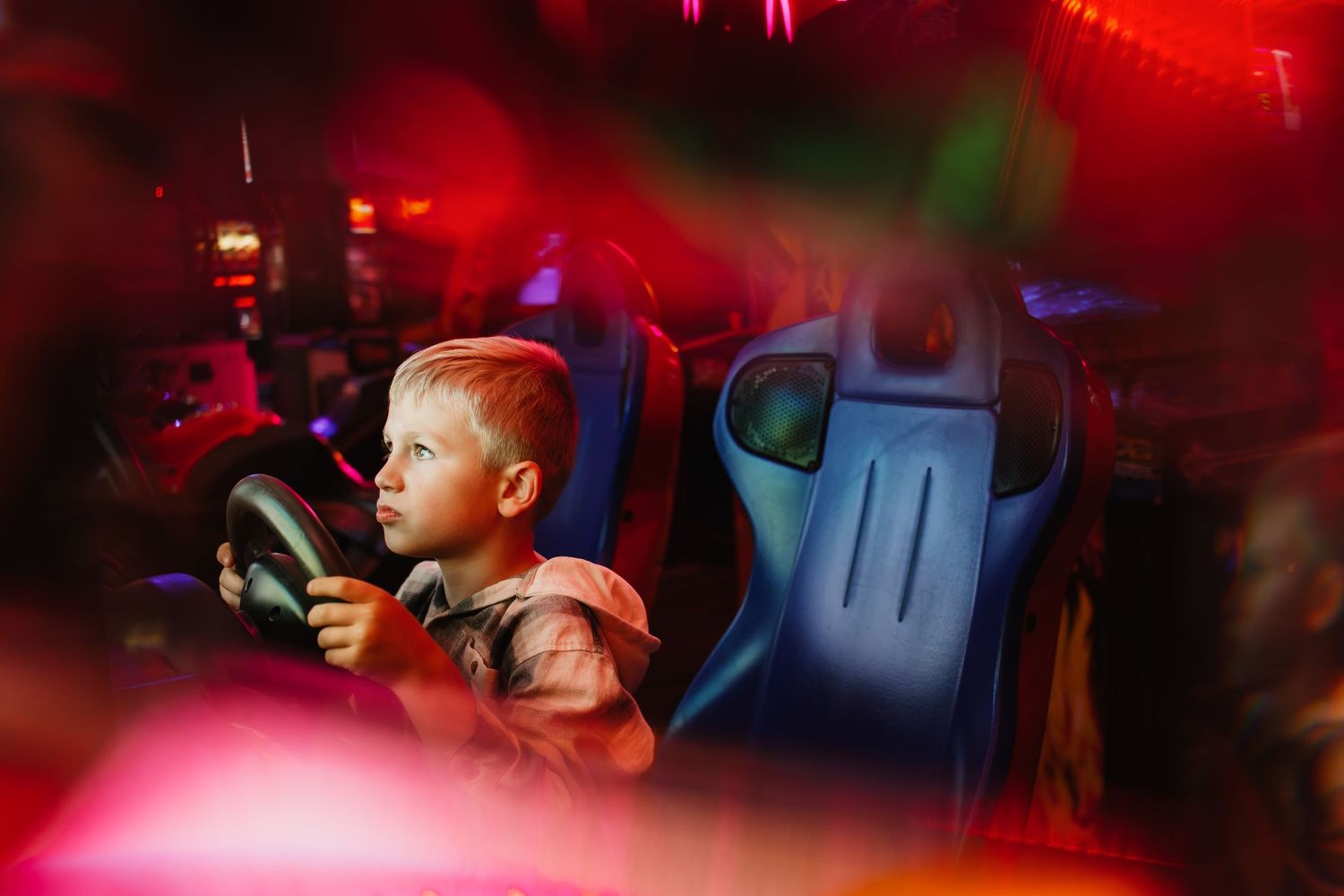 Young boy focused on steering wheel playing arcade racing game in colorful game room.