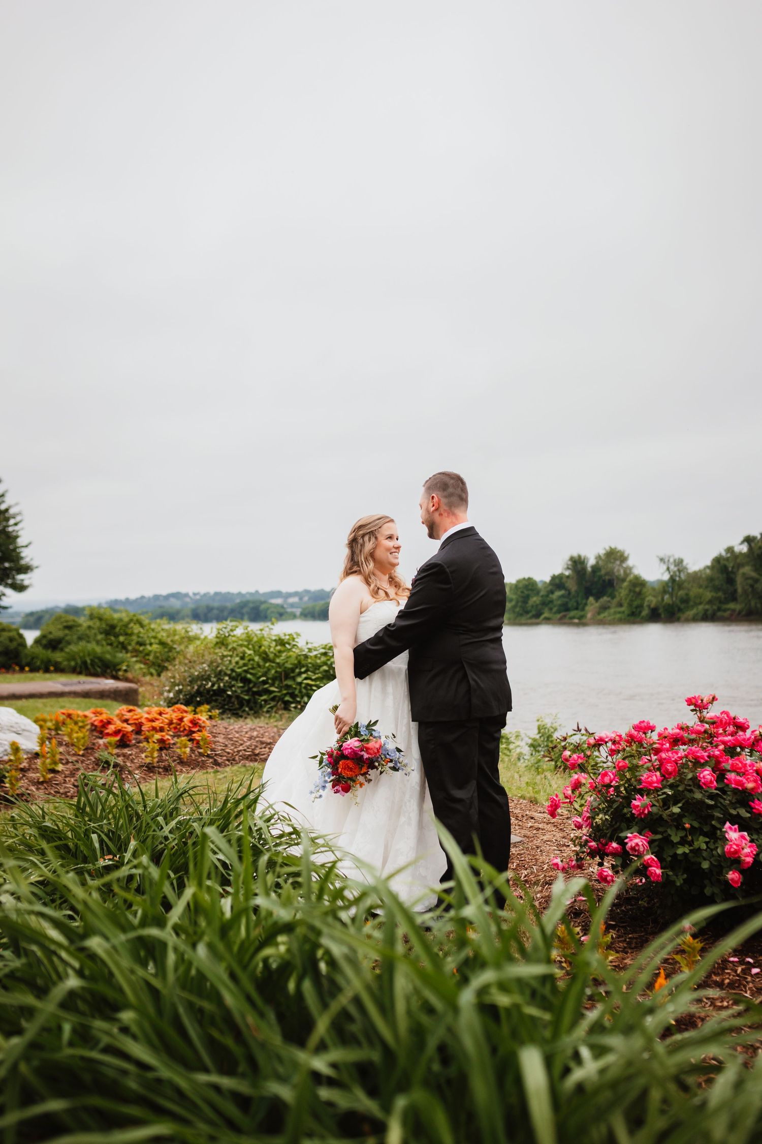 Bride holding red bouquet and groom holding hands in a lush garden with a river backdrop on wedding day.