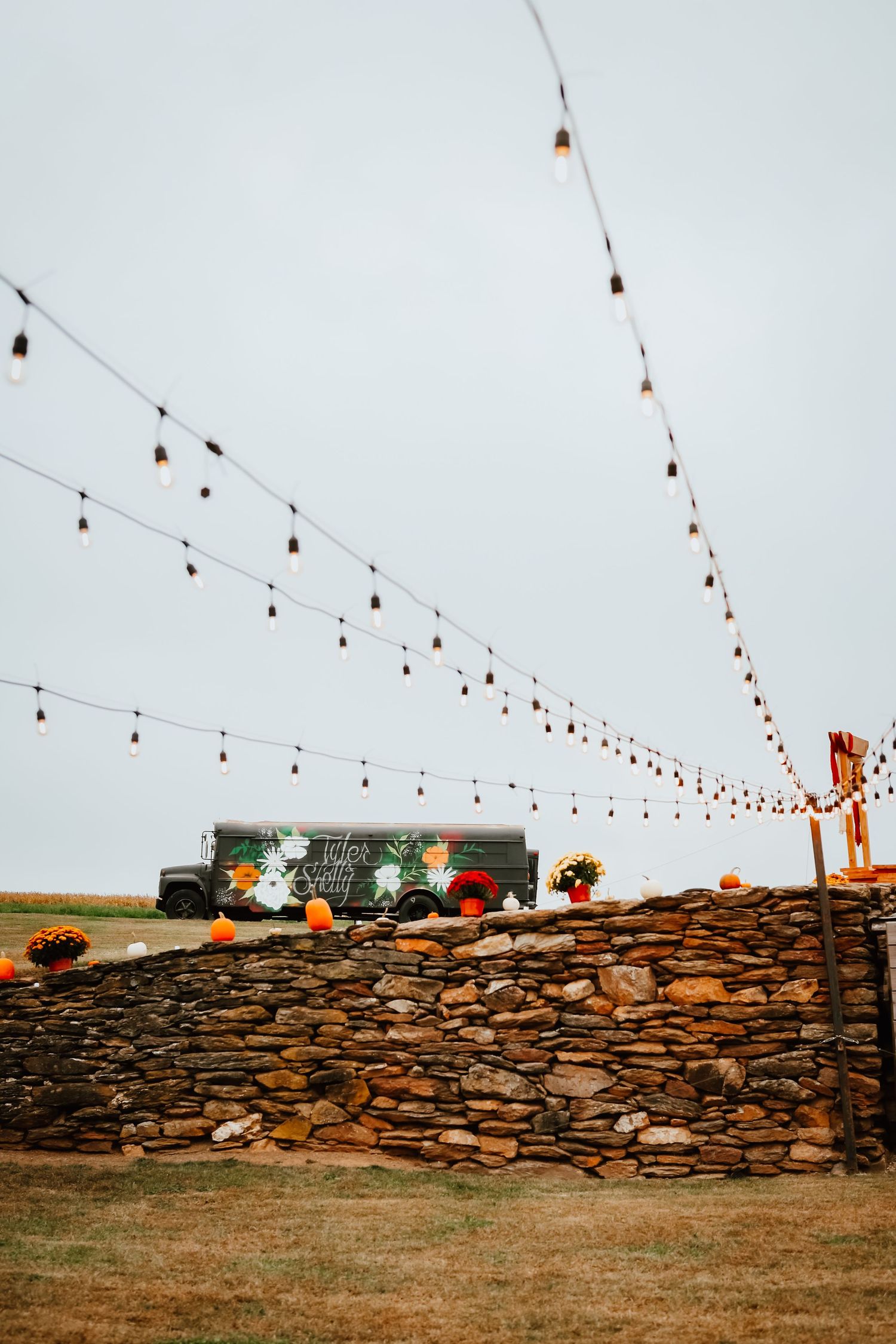 Outdoor fall wedding venue with string lights over stone wall and decorated food truck backdrop.